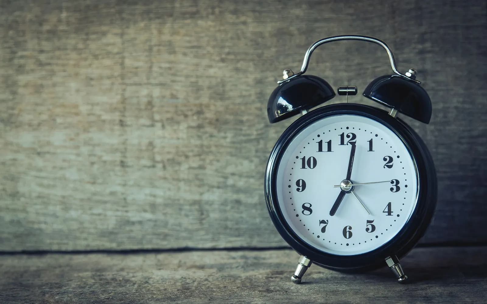 A black twin-bell alarm clock showing about 7 o'clock, on a weathered wooden surface.