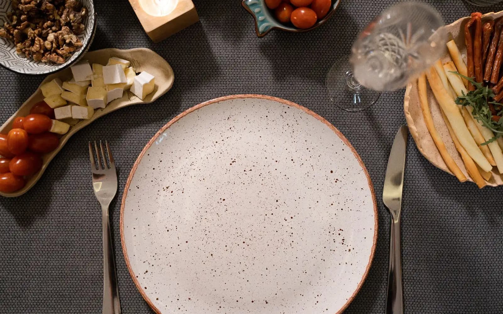 An empty plate with a fork and knife, surrounded by bowls of cheese, tomatoes and walnuts on a dark tablecloth.
