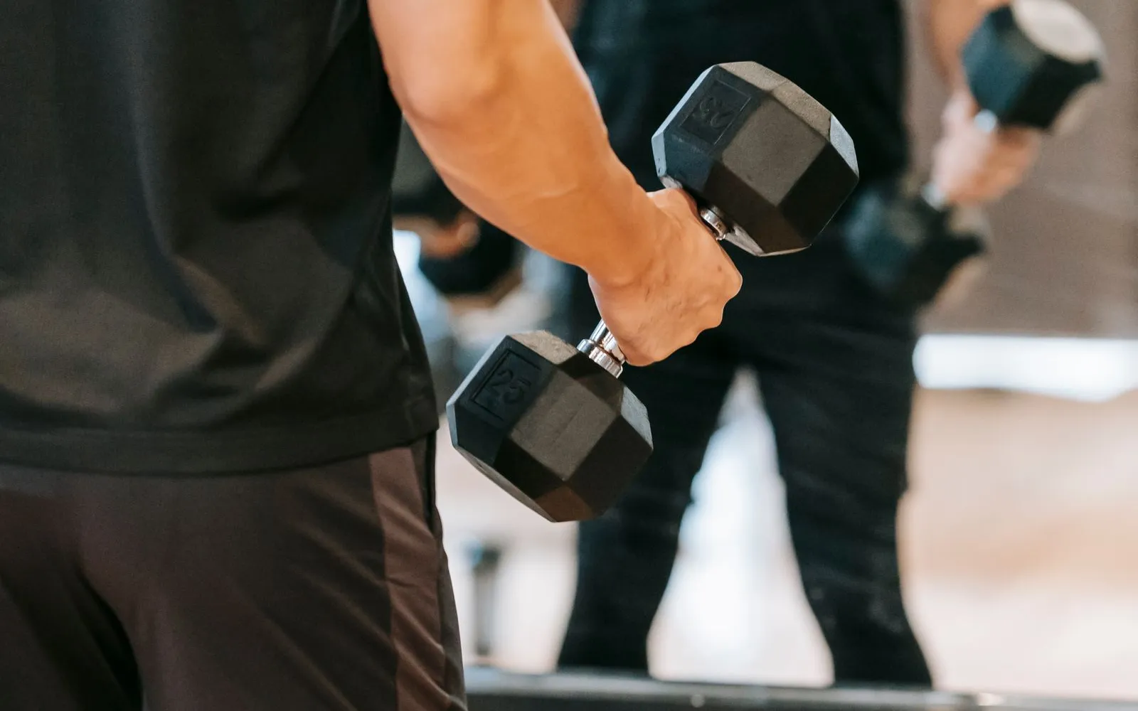 A person's arm holding a black hexagonal dumbbell during a curl, with a gym mirror behind