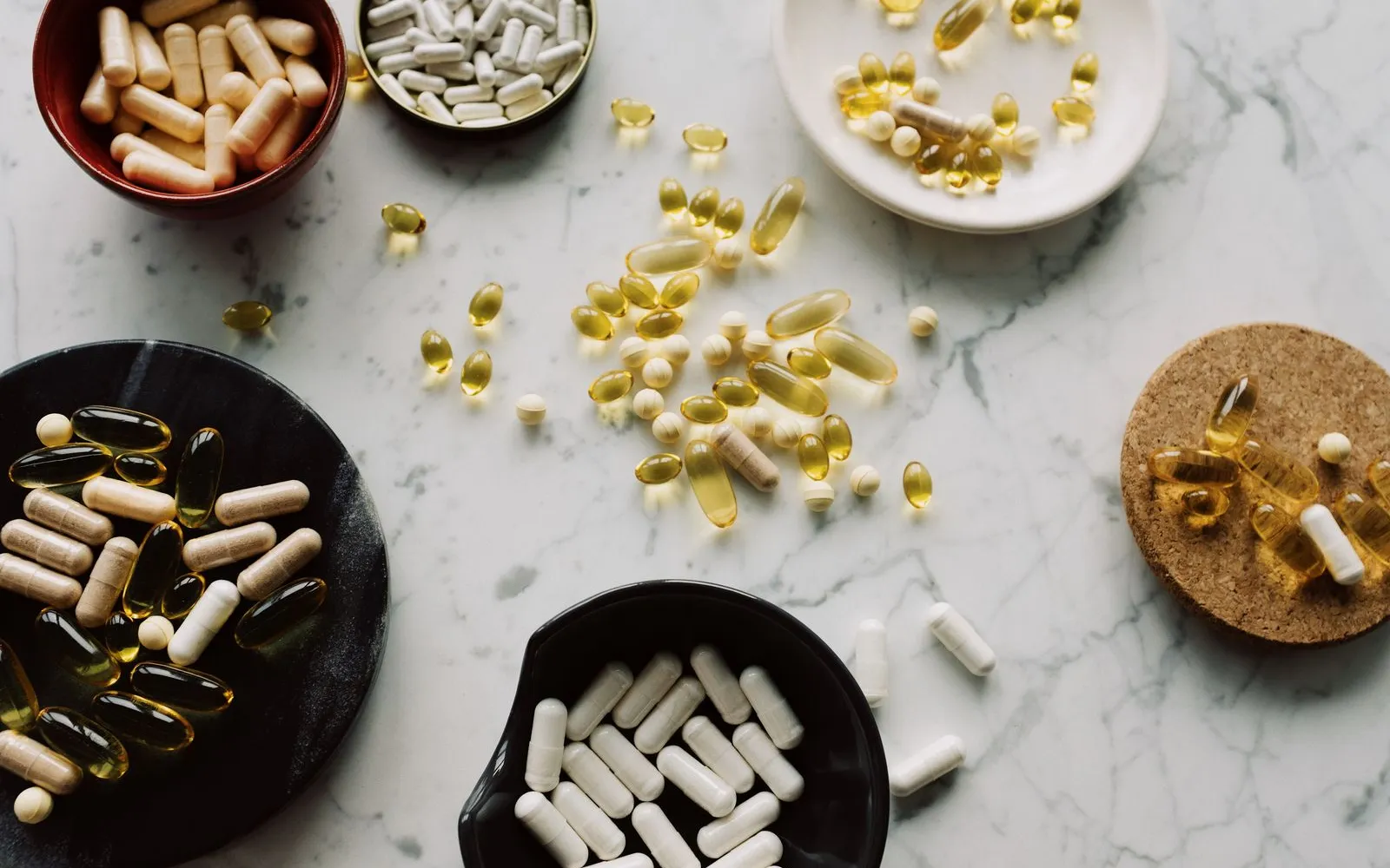 Fish oil capsules and other supplement capsules in small bowls on a marble table, seen from above