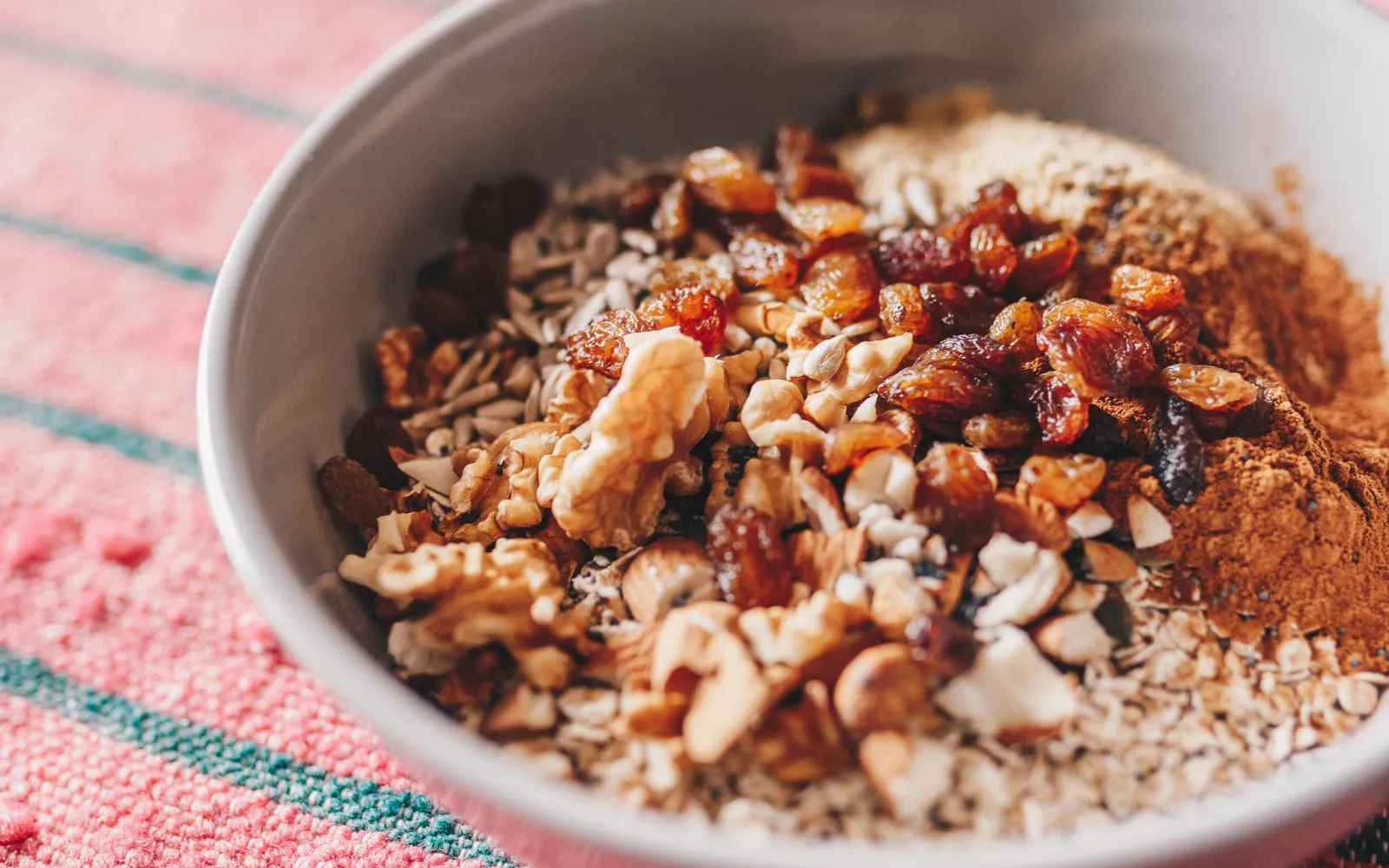 A grey bowl of rolled oats topped with walnuts, chopped almonds, sunflower seeds, raisins and cinnamon on a pink striped cloth