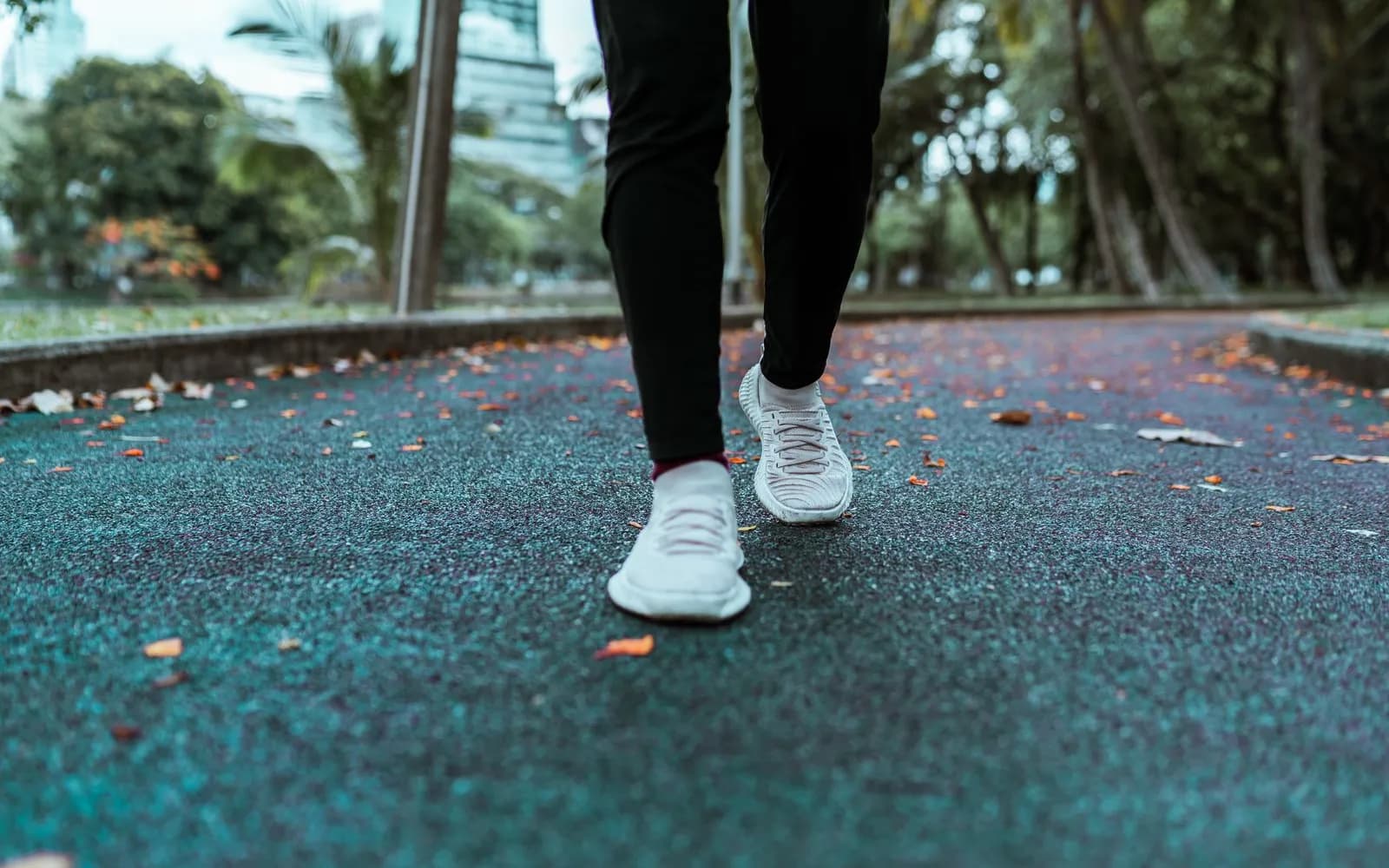 Legs of a person in black leggings and plain white trainers jogging slowly along a park path scattered with fallen leaves