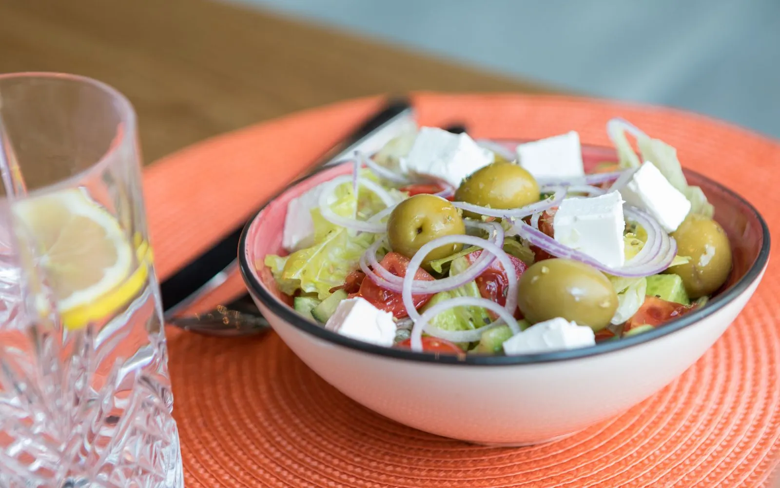 A bowl of salad with green olives, tomato, cucumber, red onion and feta, beside a glass of water with lemon