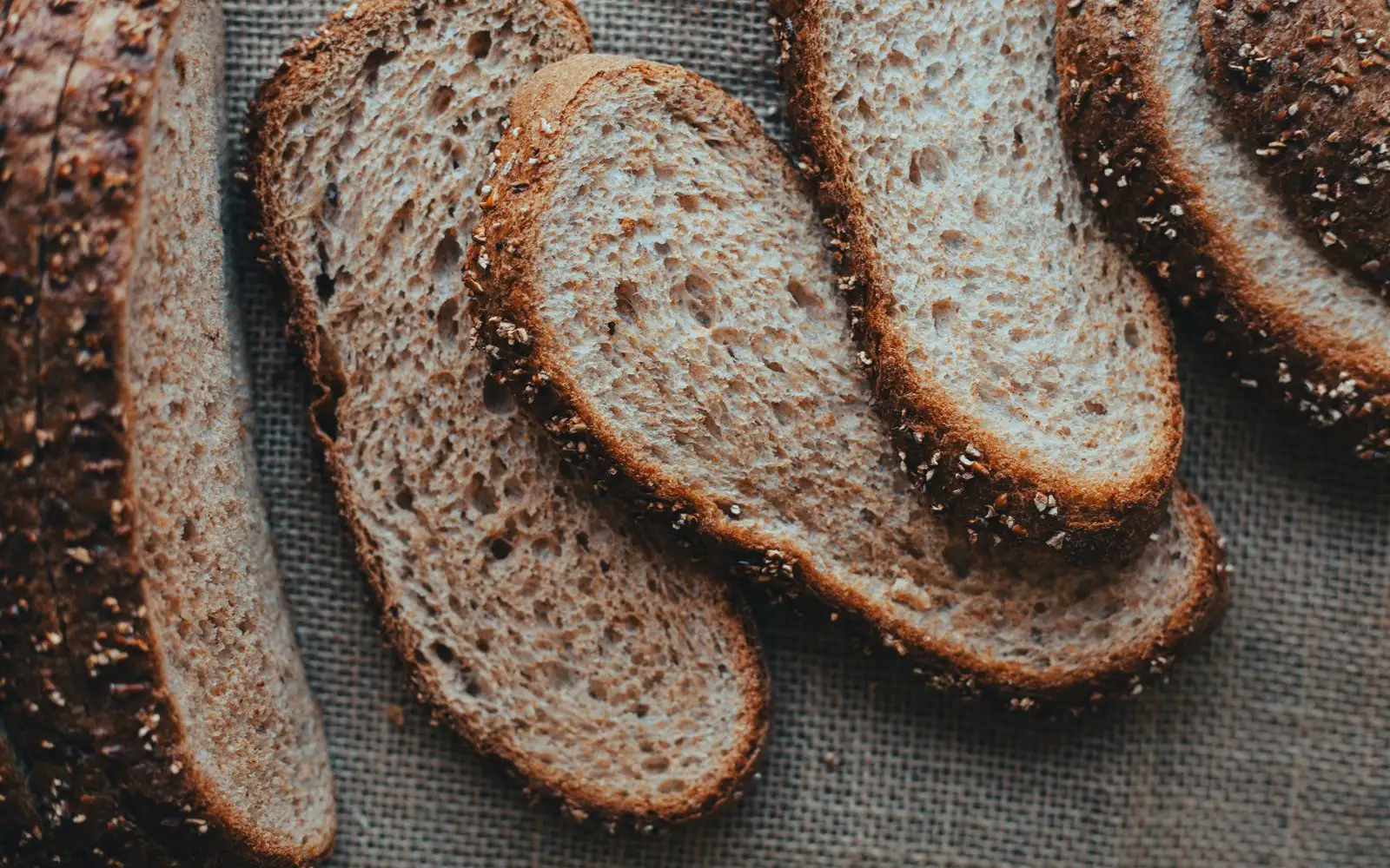 Slices of brown wholegrain bread with oat-topped crusts laid on a hessian cloth