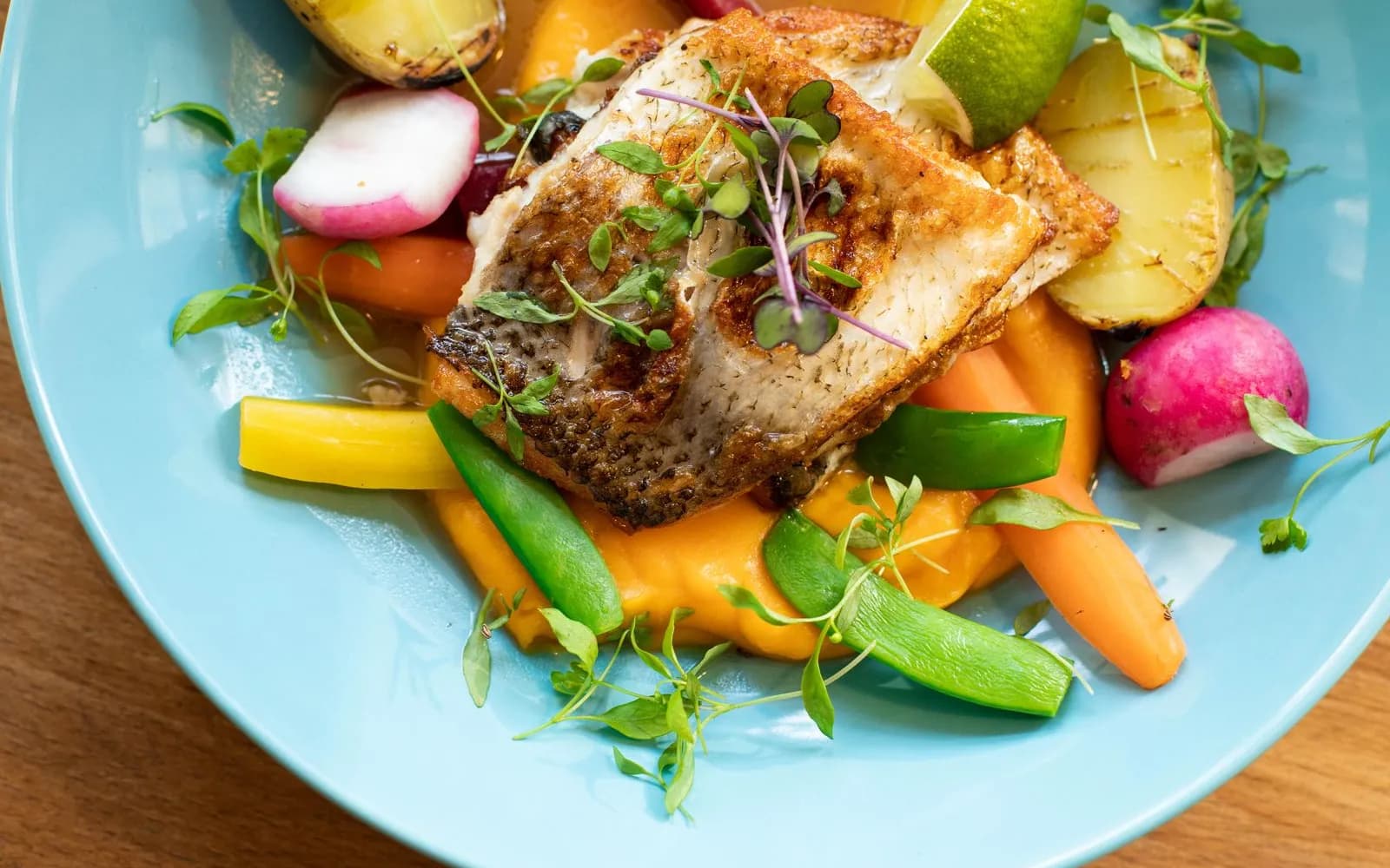 A pan-fried fish fillet on a blue plate with carrots, sugar snap peas, radishes, new potatoes and a lime wedge