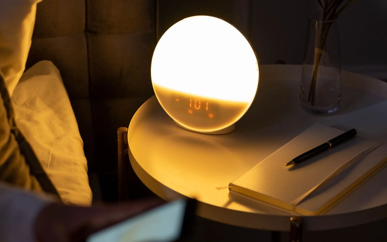 A glowing round bedside clock on a small white table next to a bed at night, with a notebook and pen.