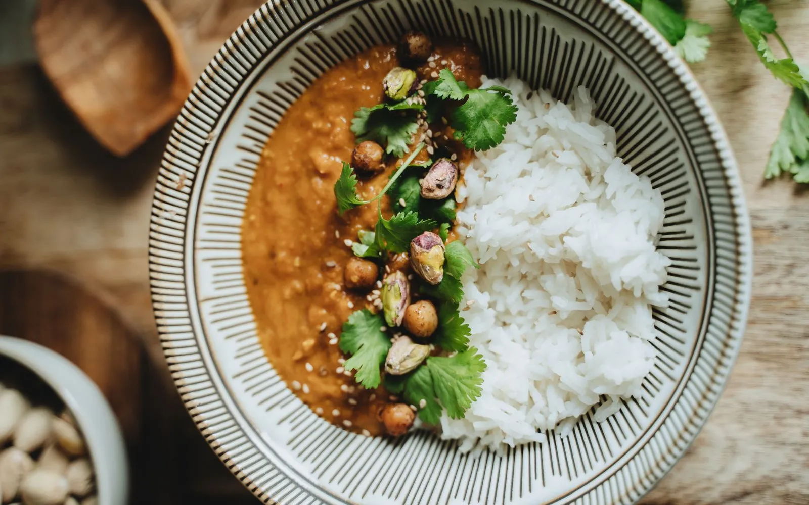 A bowl of lentil curry topped with roasted chickpeas, pistachios and coriander, beside white rice, on a wooden table