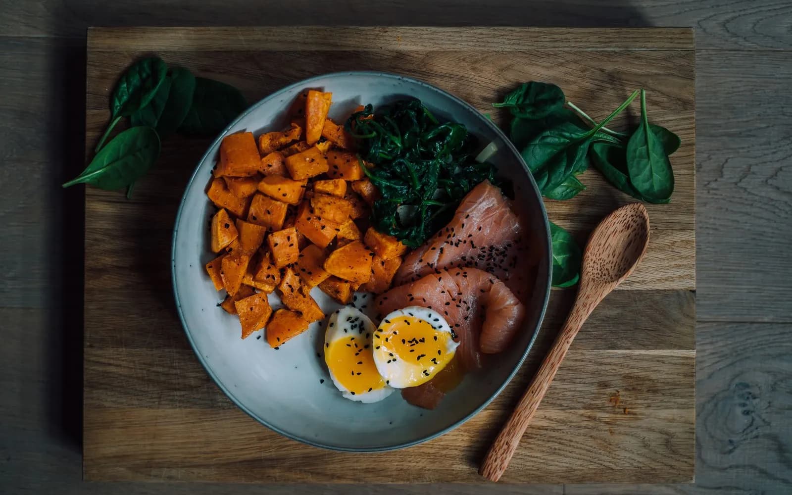 A bowl of smoked salmon, a halved soft-boiled egg, roasted sweet potato and wilted spinach on a wooden board