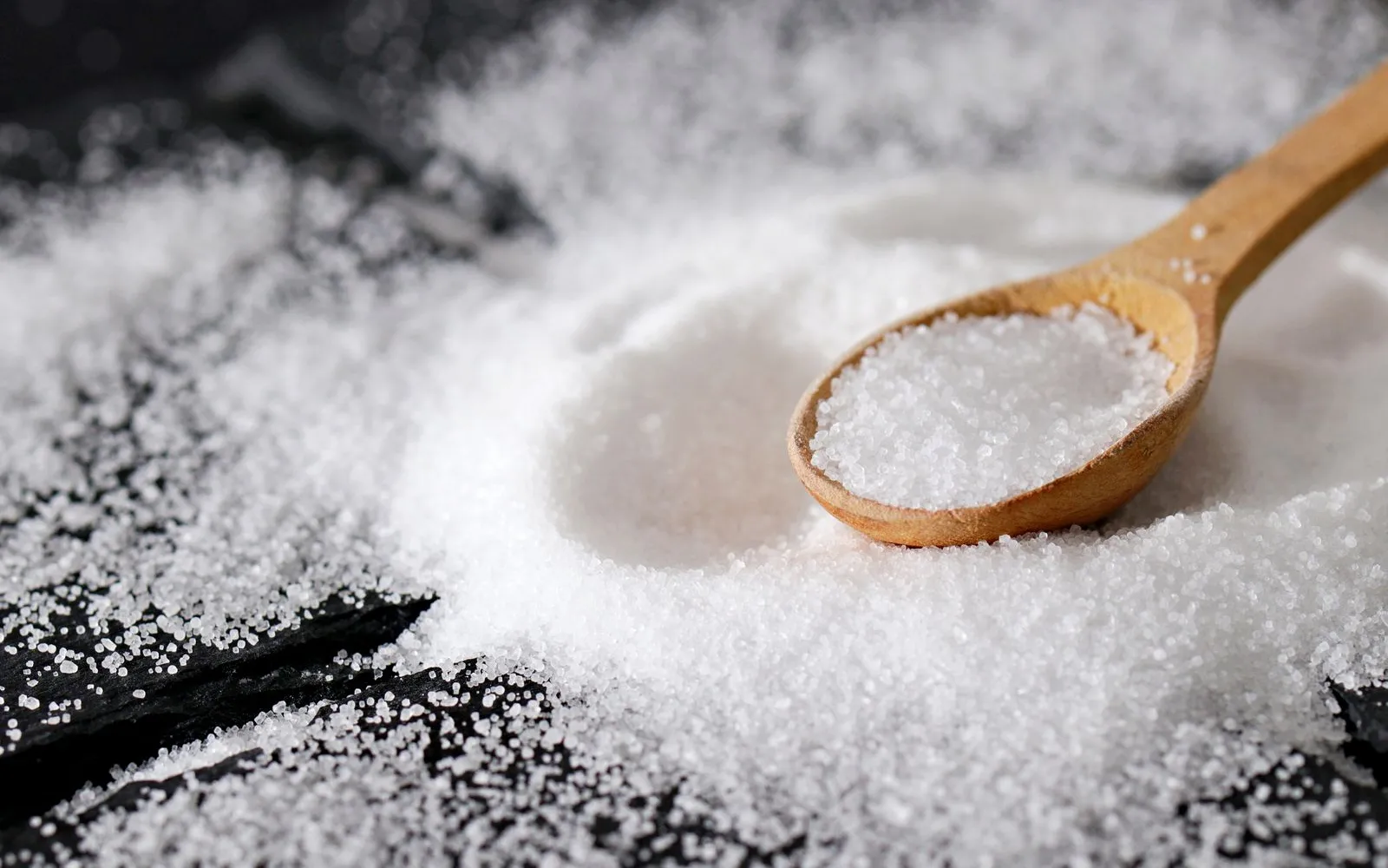 A wooden spoon filled with fine white salt, resting in a spread of salt on a dark slate surface