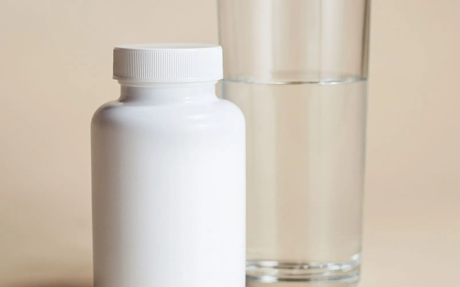 A plain white supplement tub with no label, a glass of water and three capsules on a beige surface.