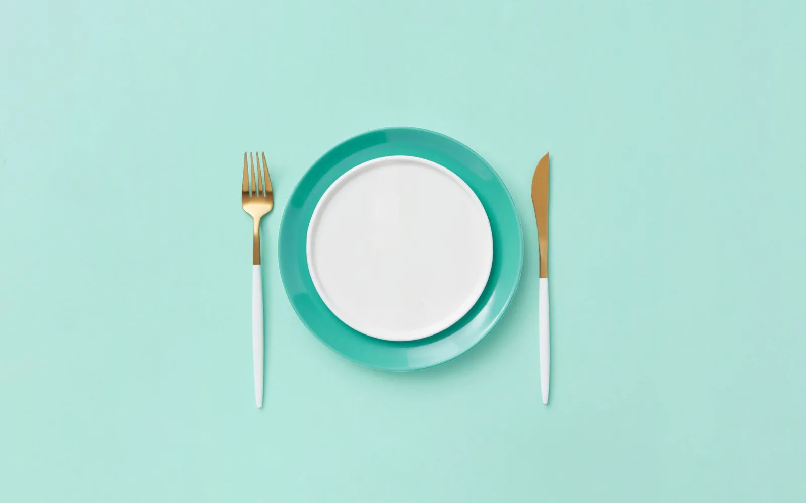 An empty white plate on a teal plate, with a gold fork and knife either side, on a teal background