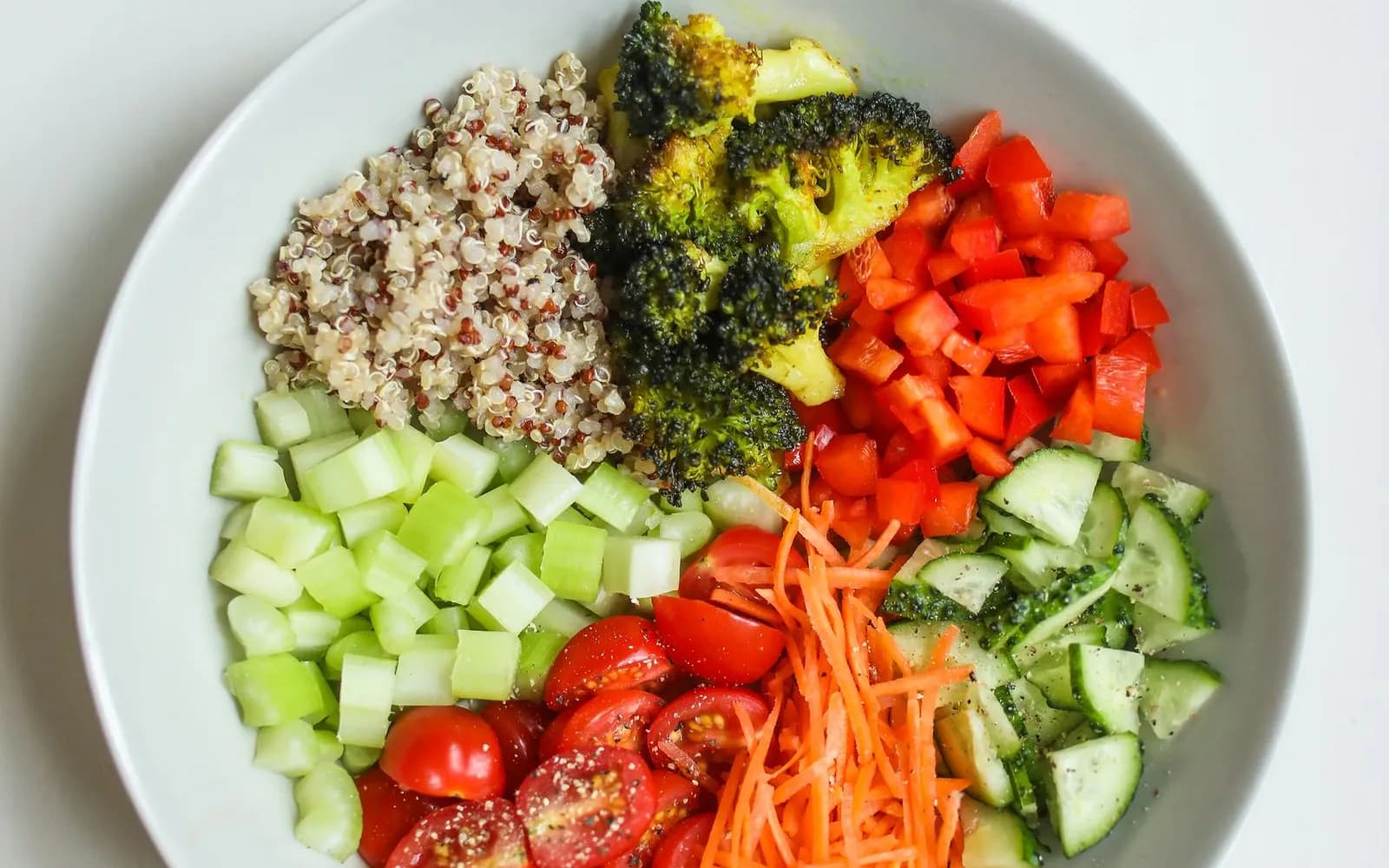 A white bowl of quinoa, roasted broccoli, red pepper, cucumber, cherry tomatoes, celery and grated carrot, seen from above