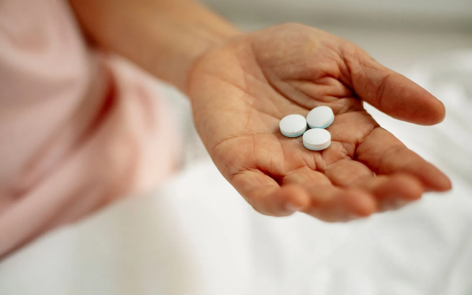 An open hand holding three small white tablets above white bedding