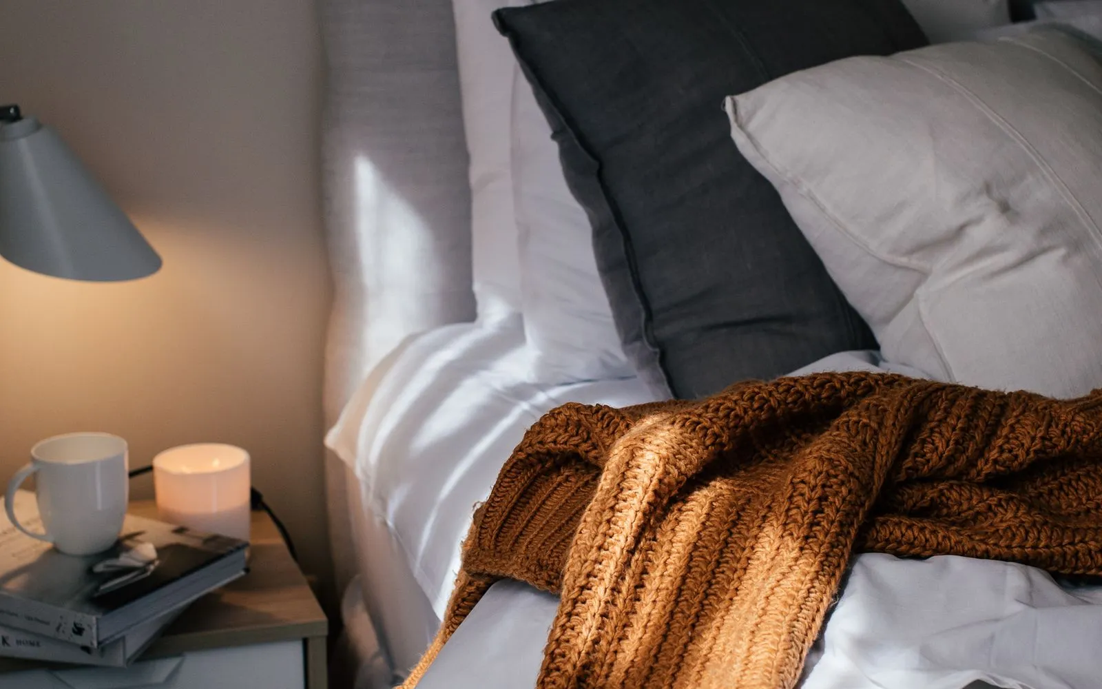 A bedside table with a lit lamp, a candle, a mug and books beside a bed with grey pillows and a rust-coloured knitted throw.