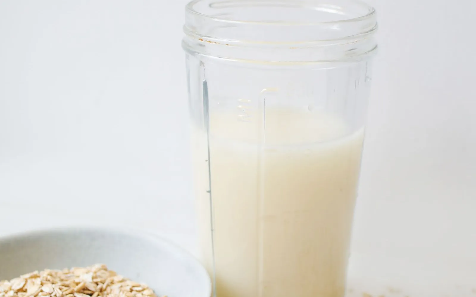 A glass jar of oat milk beside a bowl of rolled oats, with loose oats scattered on a white table