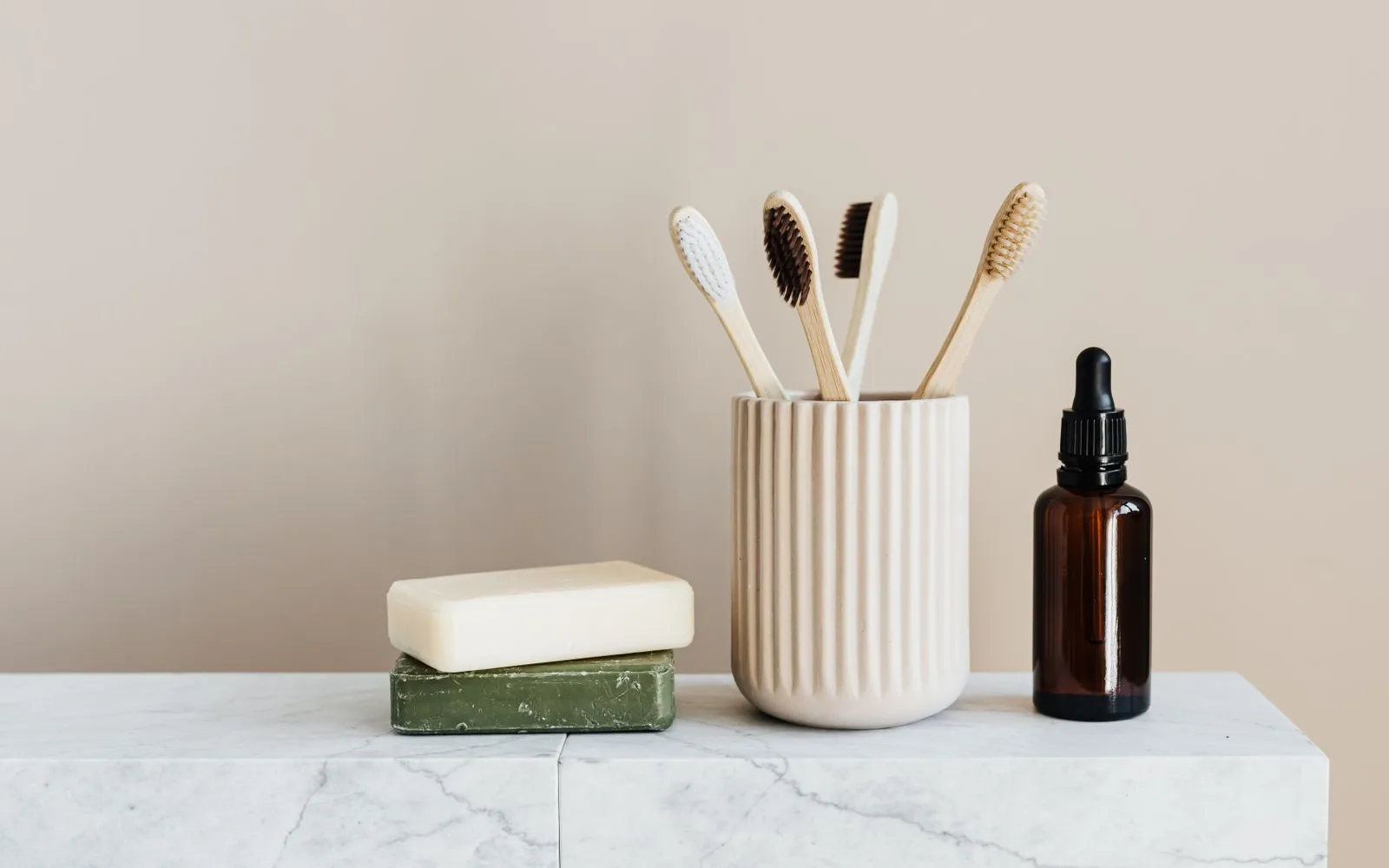 Four bamboo toothbrushes in a ribbed cup beside two bars of soap and a brown glass dropper bottle on a marble shelf