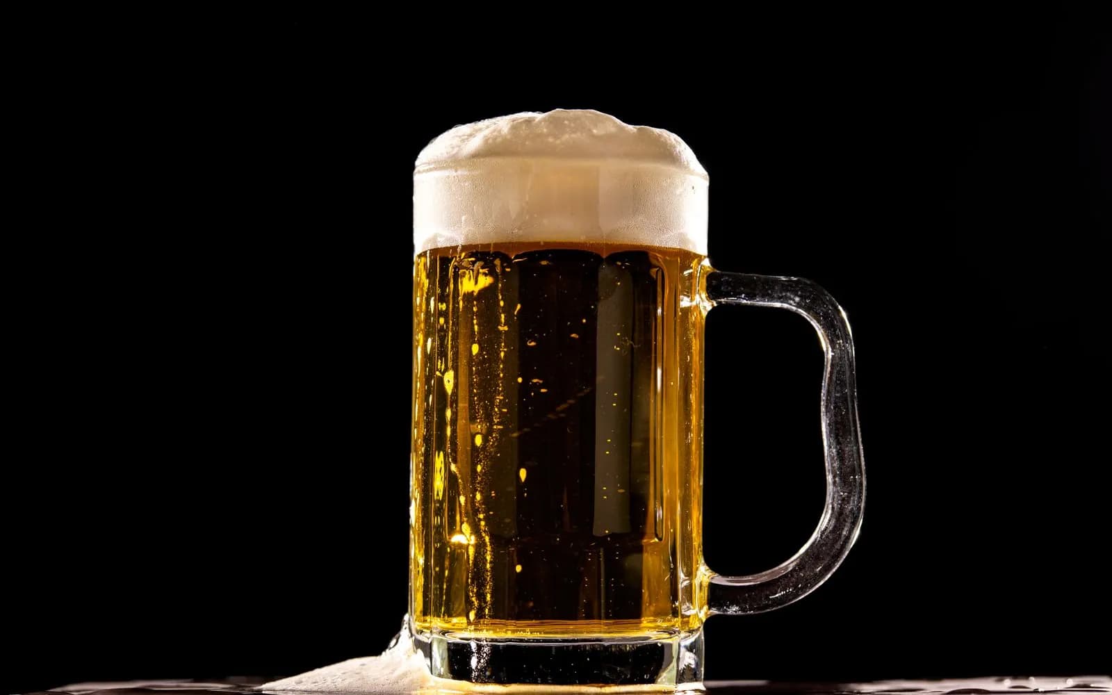 A glass mug of pale beer with a thick head of foam on a dark background.