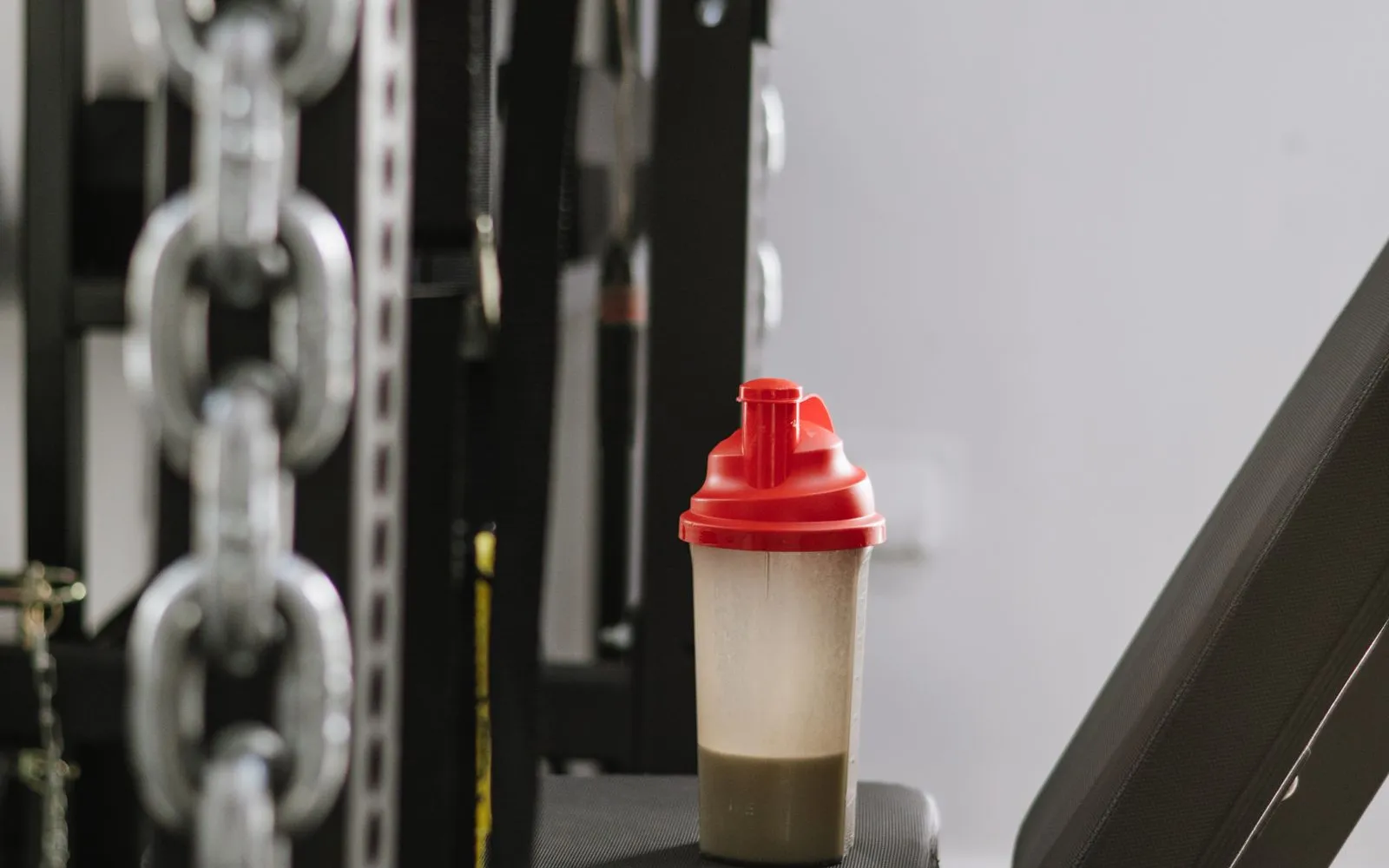 A protein shake in a plastic shaker bottle with a red lid, standing on a weight bench in a gym