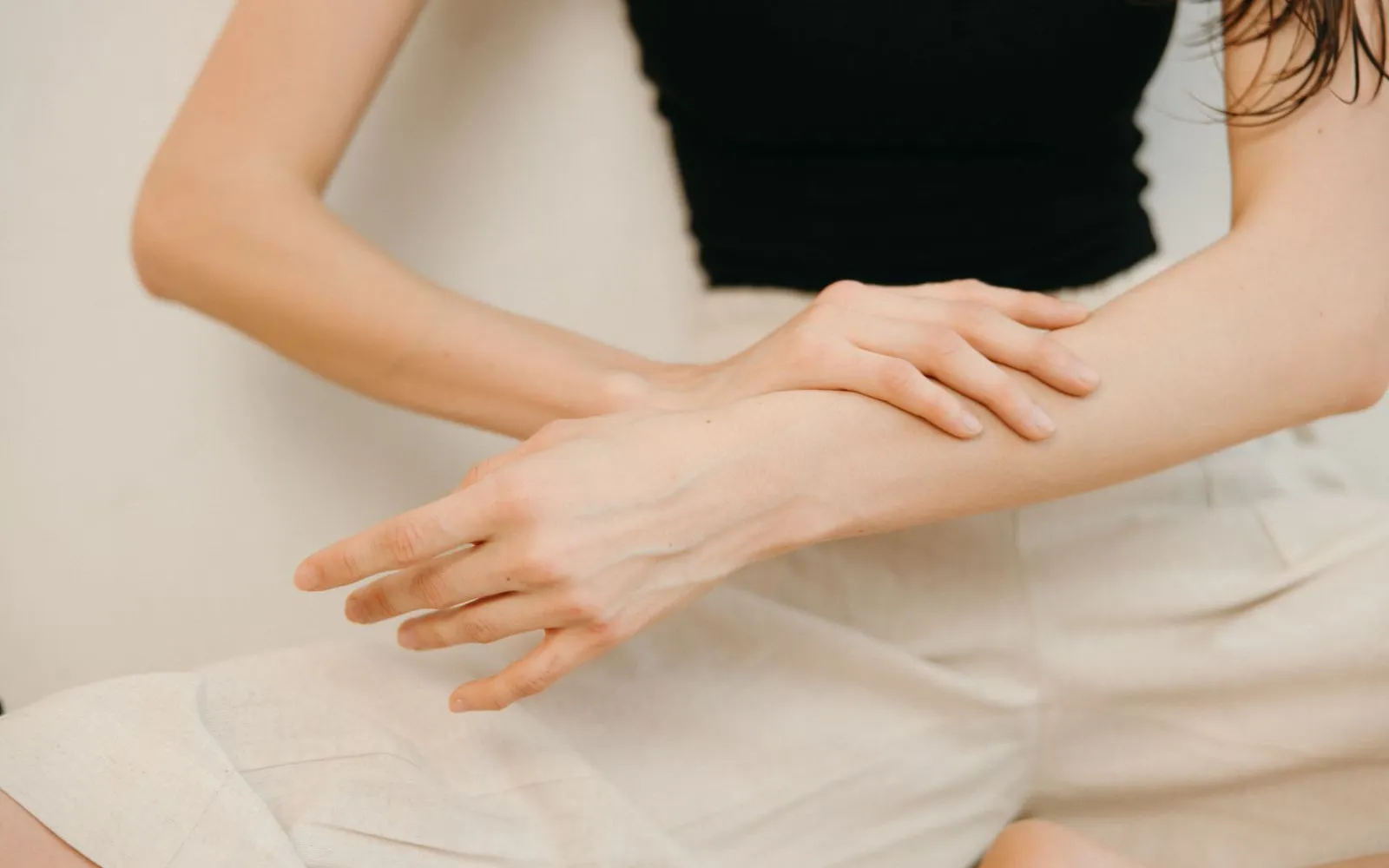 One hand gently rubbing the forearm of the other arm, against a plain cream background.