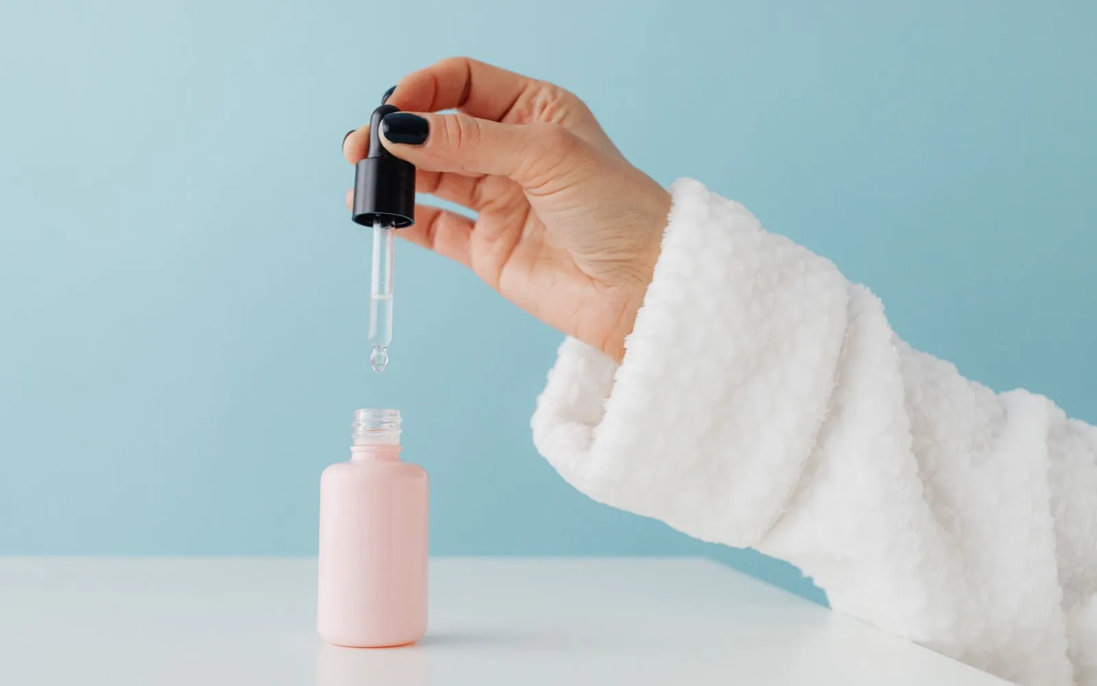 A hand in a white towelling sleeve lets a drop of clear serum fall from a dropper into a plain pink bottle on a white table