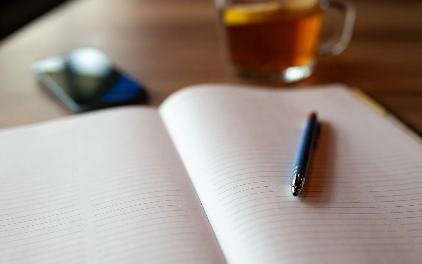 An open lined notebook with a pen on the page, a glass mug of tea and a phone blurred behind it on a wooden table.