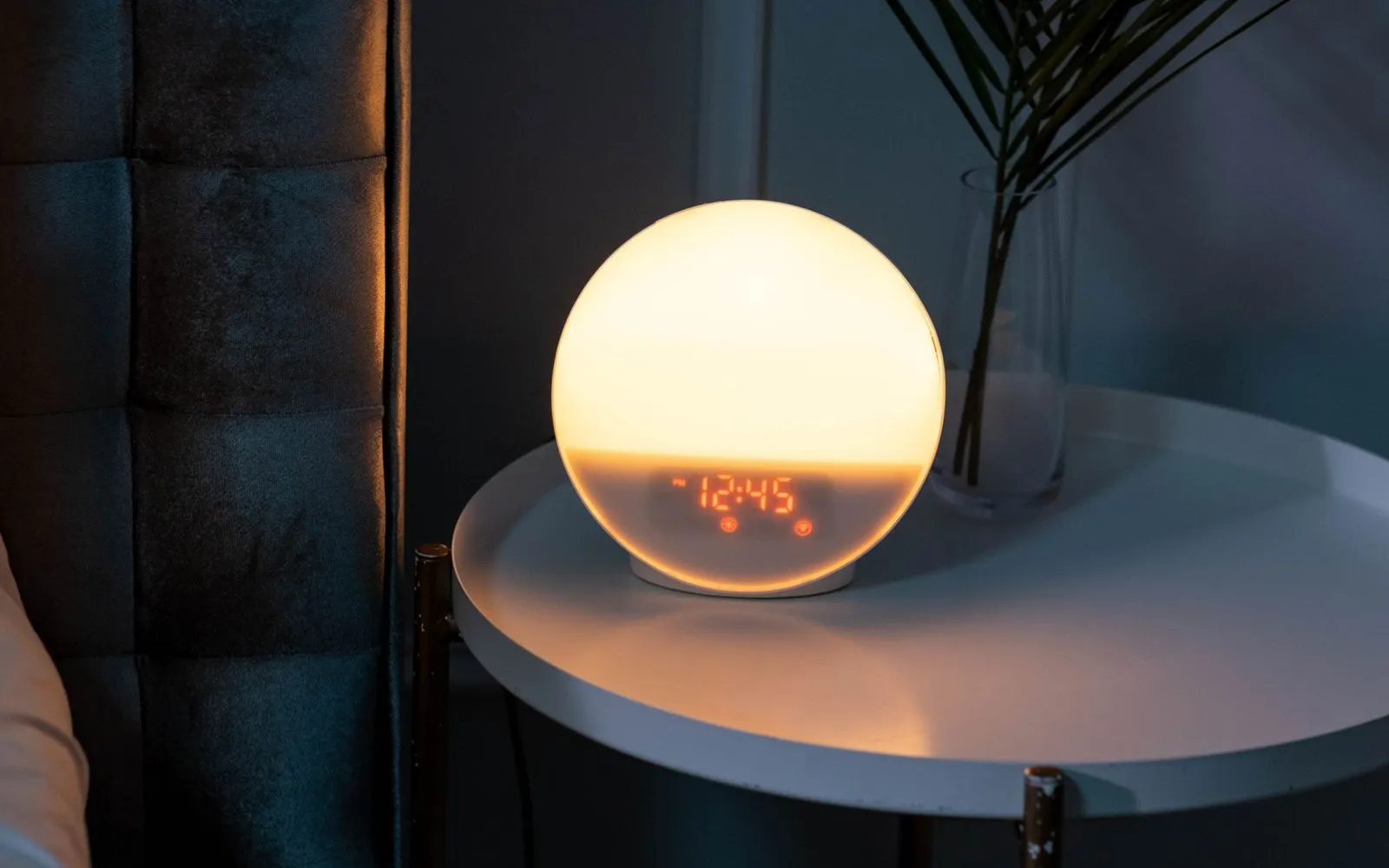 A glowing round alarm clock on a small white bedside table in a dark bedroom, next to a palm leaf in a glass vase