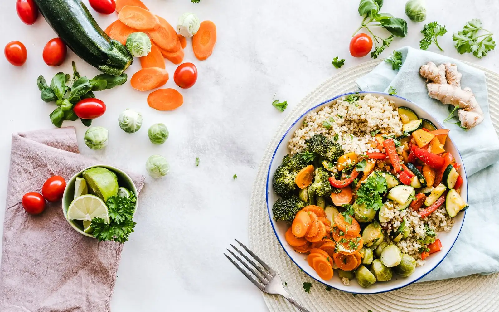 A bowl of quinoa with broccoli, carrots, peppers, courgette and sprouts, next to a fork and loose vegetables on a white table