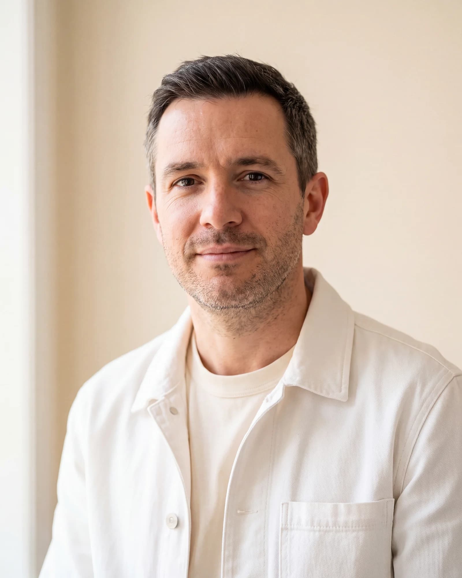 A man in his forties in a cream canvas jacket stands against a warm ivory wall, looking to camera