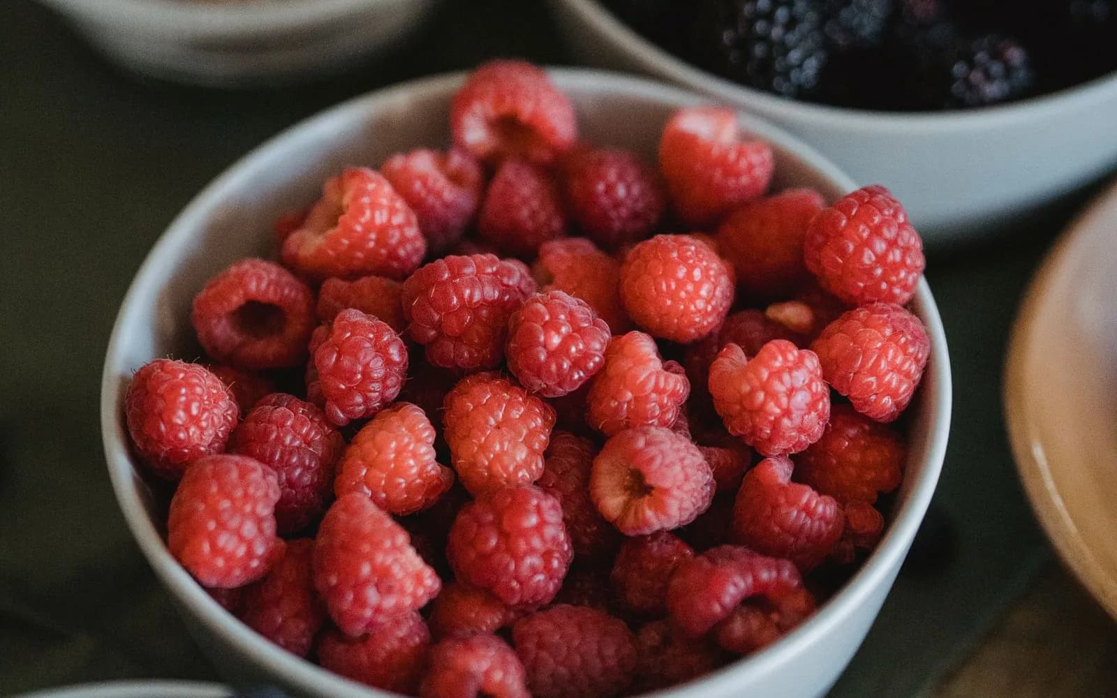 A white bowl of raspberries beside bowls of blackberries, blueberries and granola on a table