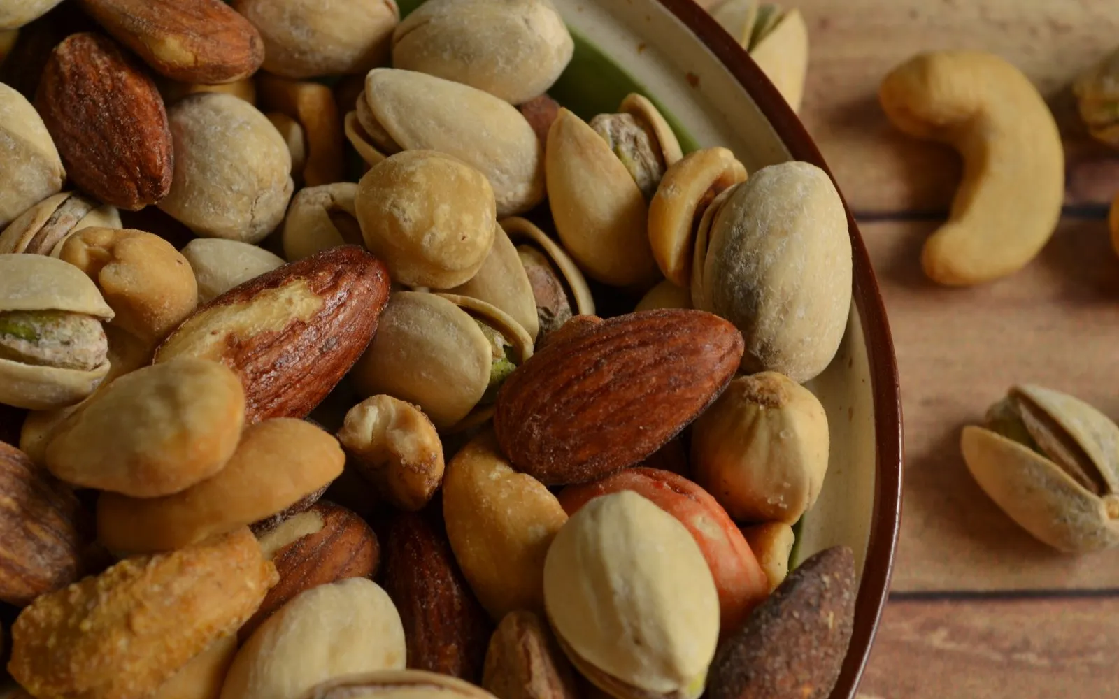 A bowl of mixed roasted nuts, including almonds, pistachios in their shells and cashews, on a wooden table