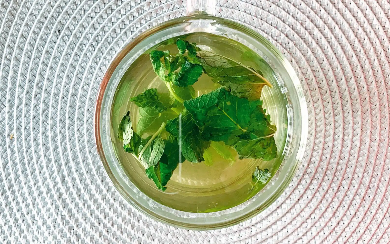 Top view of a glass cup of mint tea with fresh mint leaves on a pale woven mat