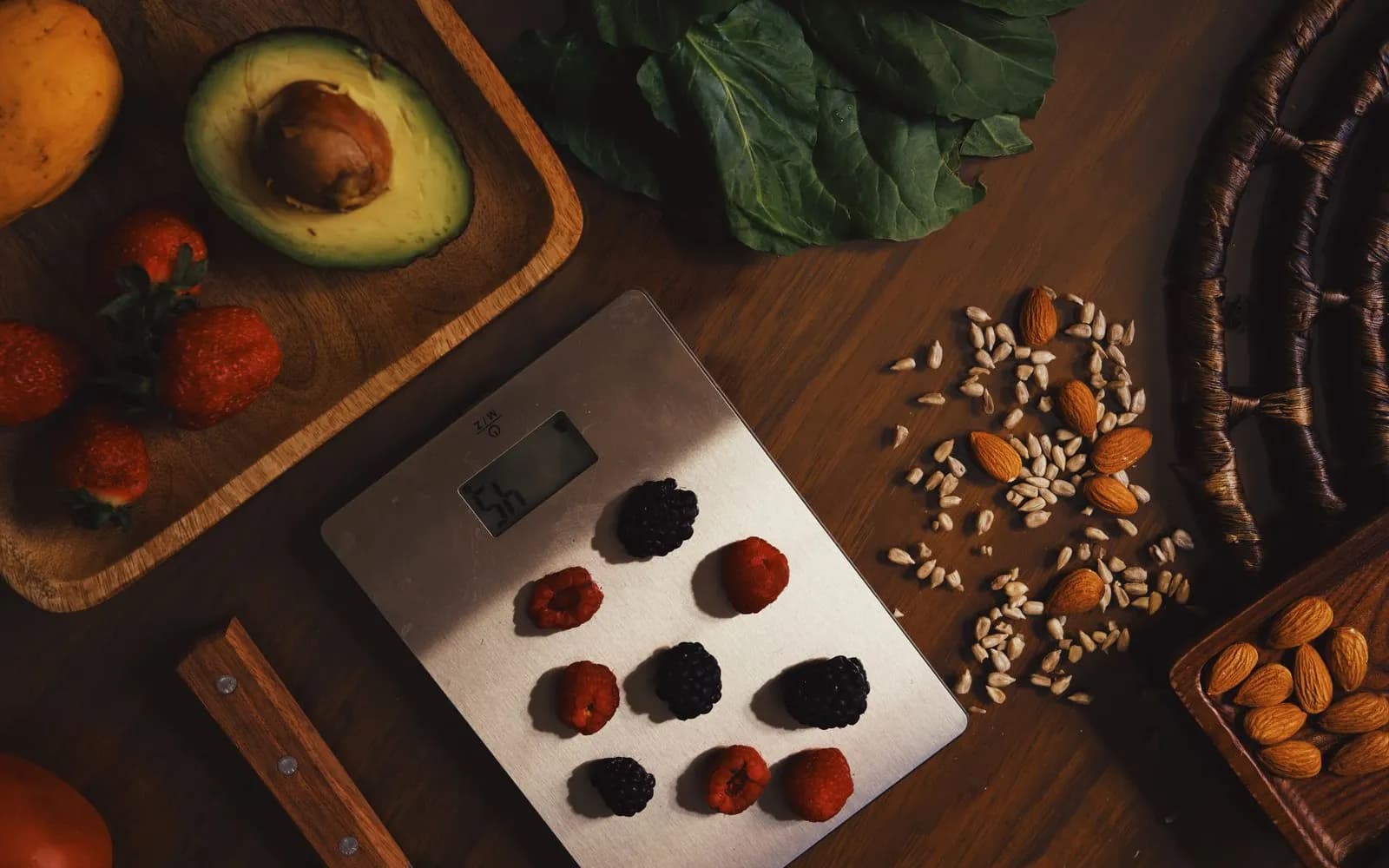 Berries on a digital kitchen scale, with almonds, seeds, avocado, strawberries and leafy greens on a wooden table