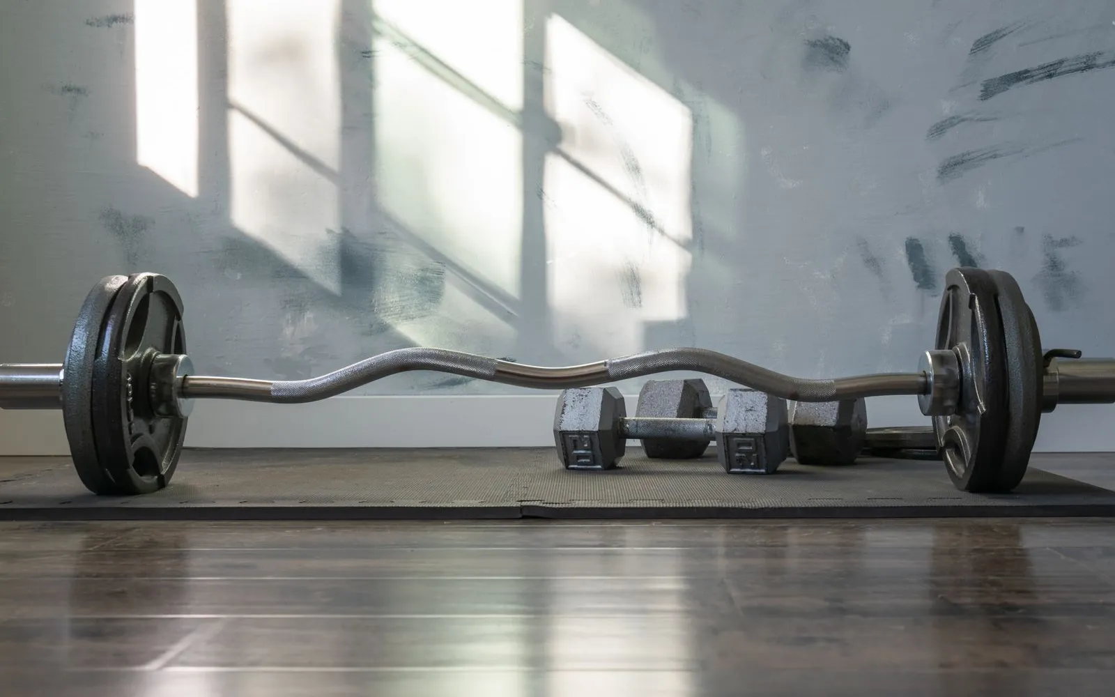 A curl barbell with weight plates and a pair of dumbbells on a gym mat, with window light on the wall behind.