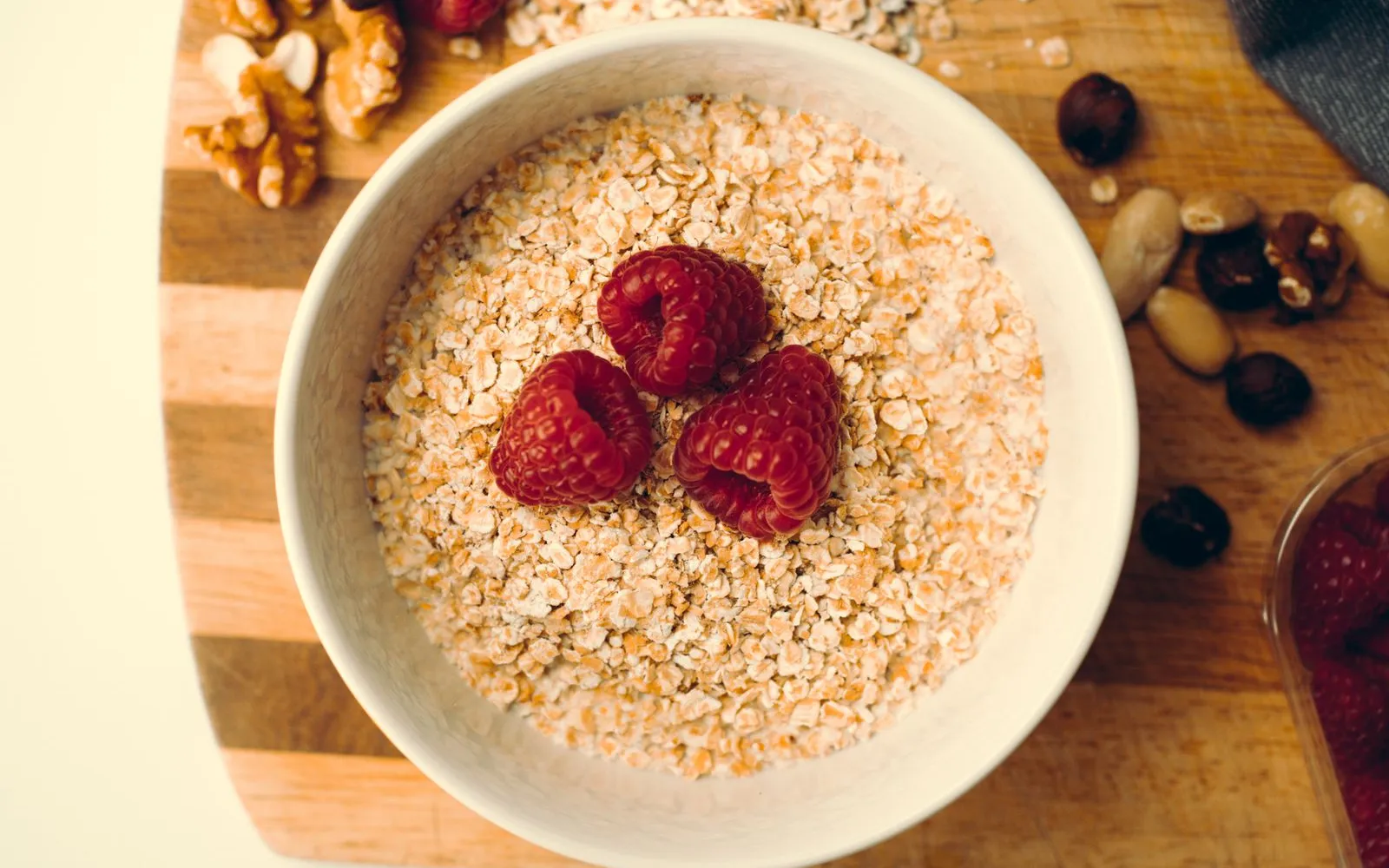 A white bowl of rolled oats topped with raspberries, with walnuts and other nuts on a wooden board