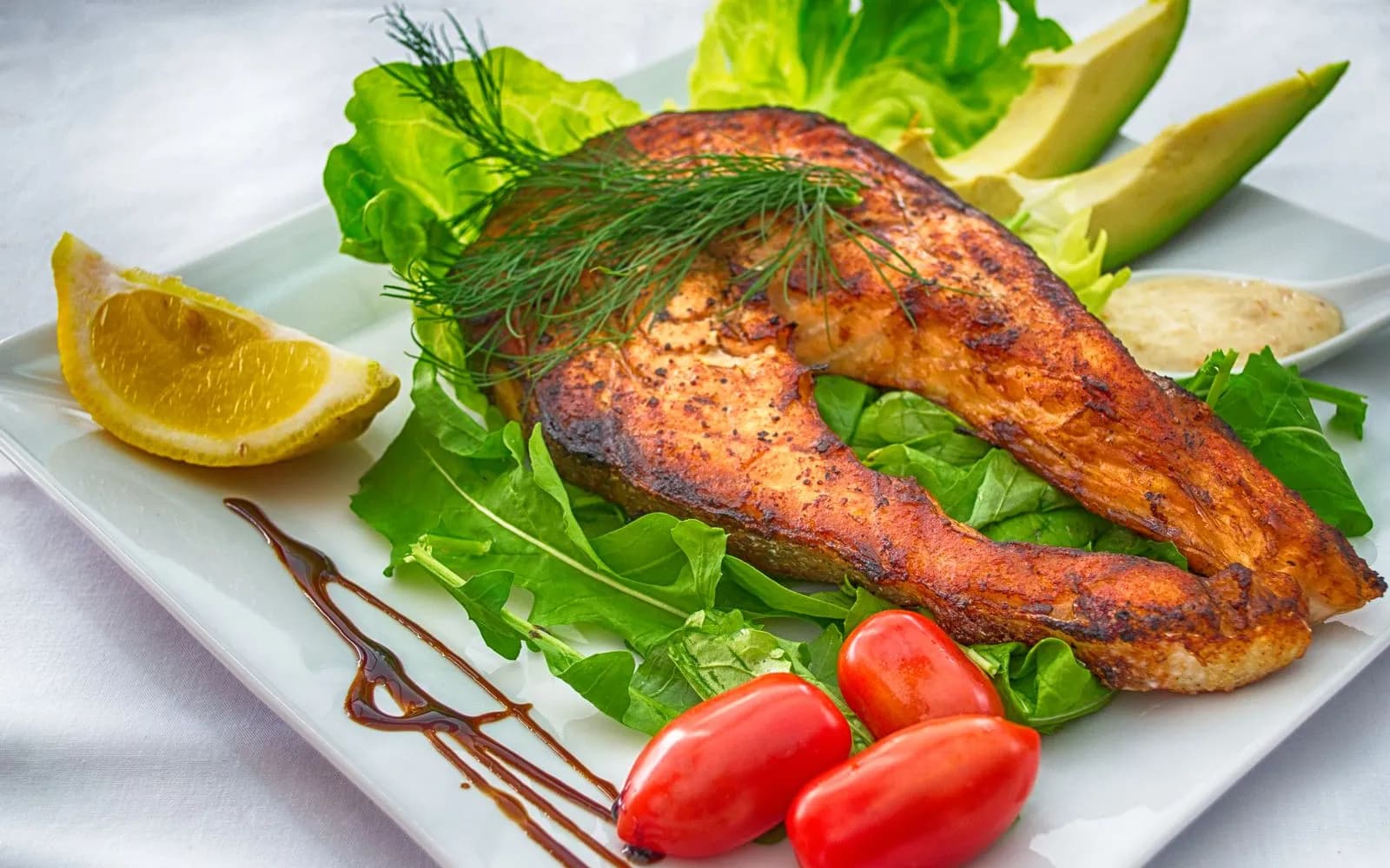 A grilled salmon steak on salad leaves with cherry tomatoes, avocado slices and a lemon wedge on a white plate