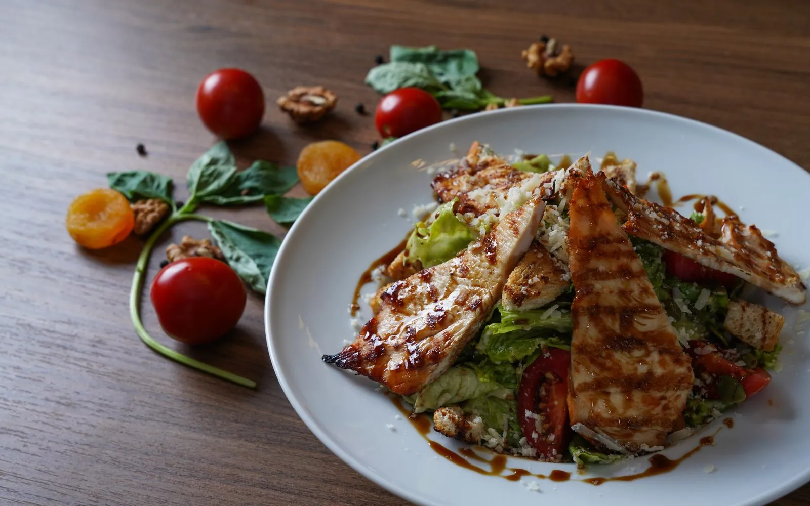 Sliced grilled chicken breast on a salad of lettuce, tomatoes, croutons and grated cheese on a white plate on a wooden table