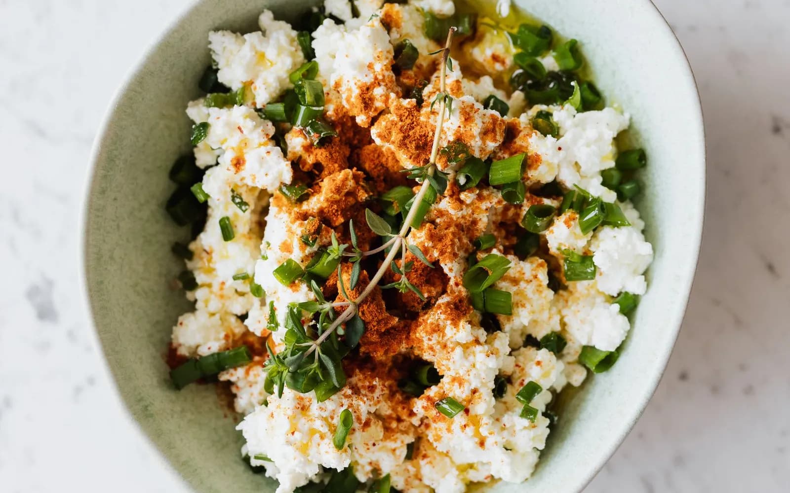 A bowl of cottage cheese topped with chopped spring onion, thyme, paprika and a little olive oil on a marble surface