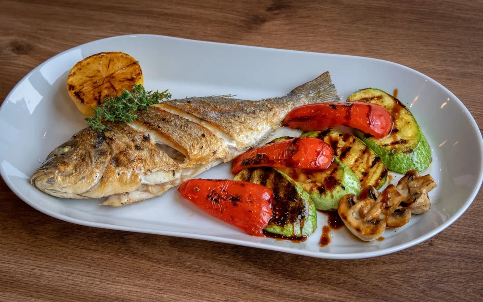 A whole pan-fried fish with grilled peppers, courgette, mushrooms and a charred lemon half on a white plate