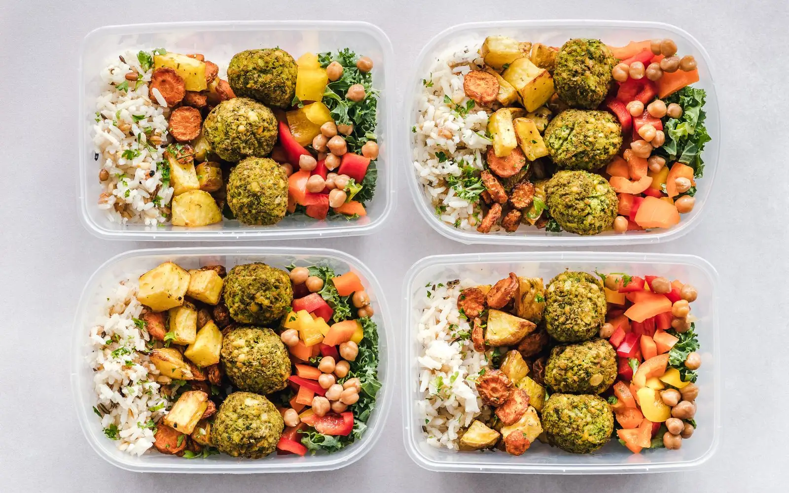 Four clear lunch boxes, each with rice, roasted vegetables, falafel, chickpeas and chopped peppers.