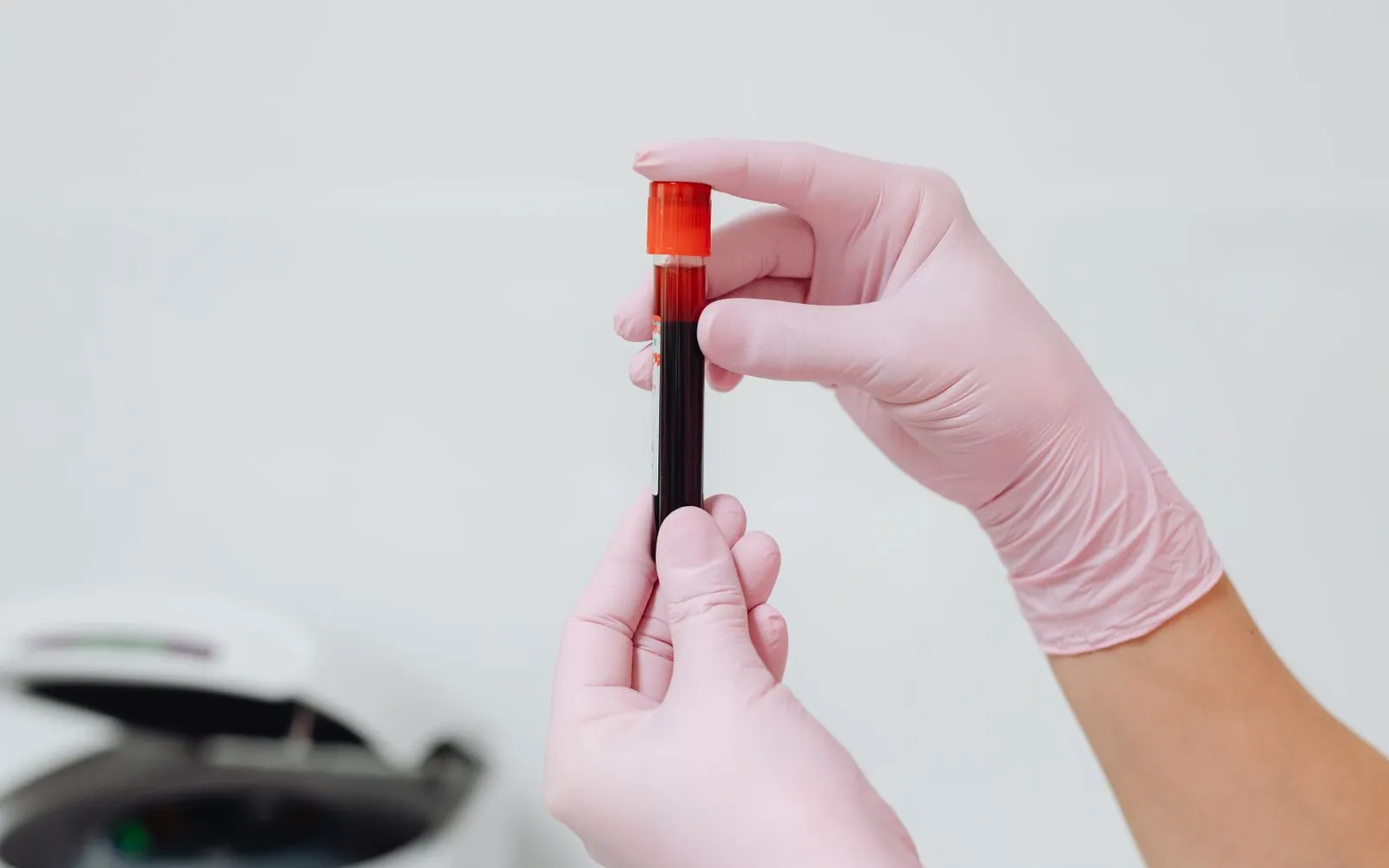 Two gloved hands holding a capped tube of blood in a lab, with a centrifuge blurred in the corner.