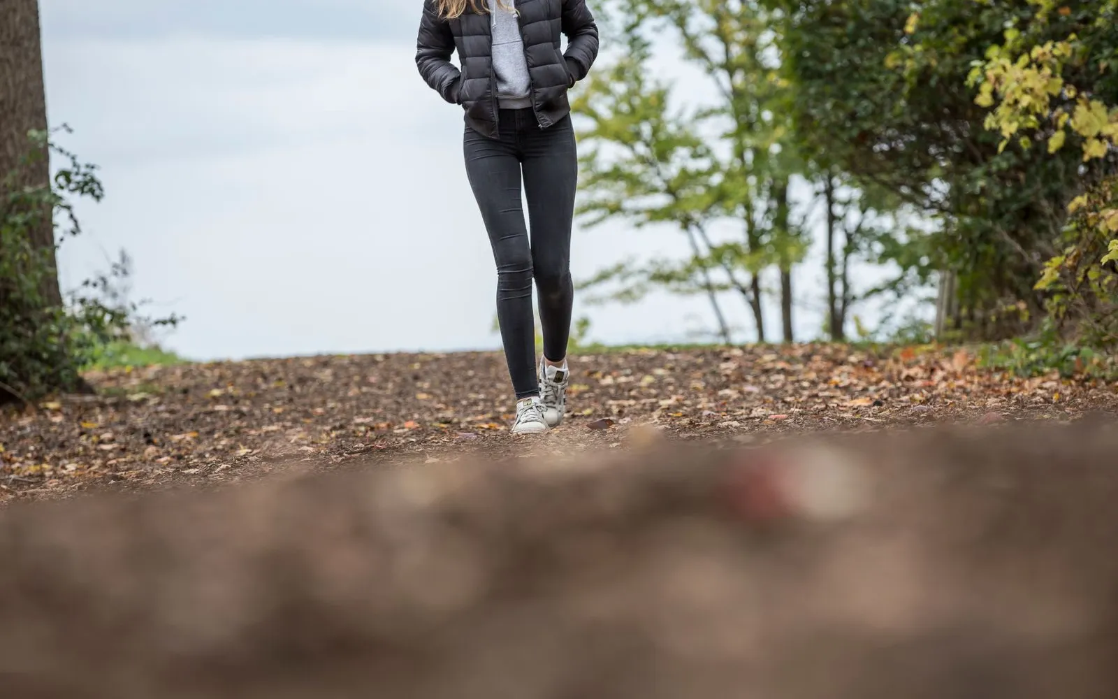 A person in a dark jacket, black leggings and white trainers walking along a leaf-covered woodland path, cropped below the face