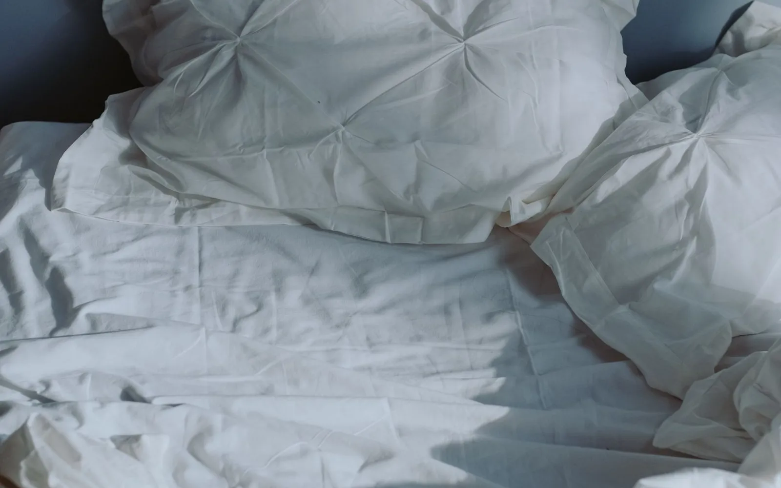 An empty, unmade bed with a white pillow and creased white sheets against a blue wall