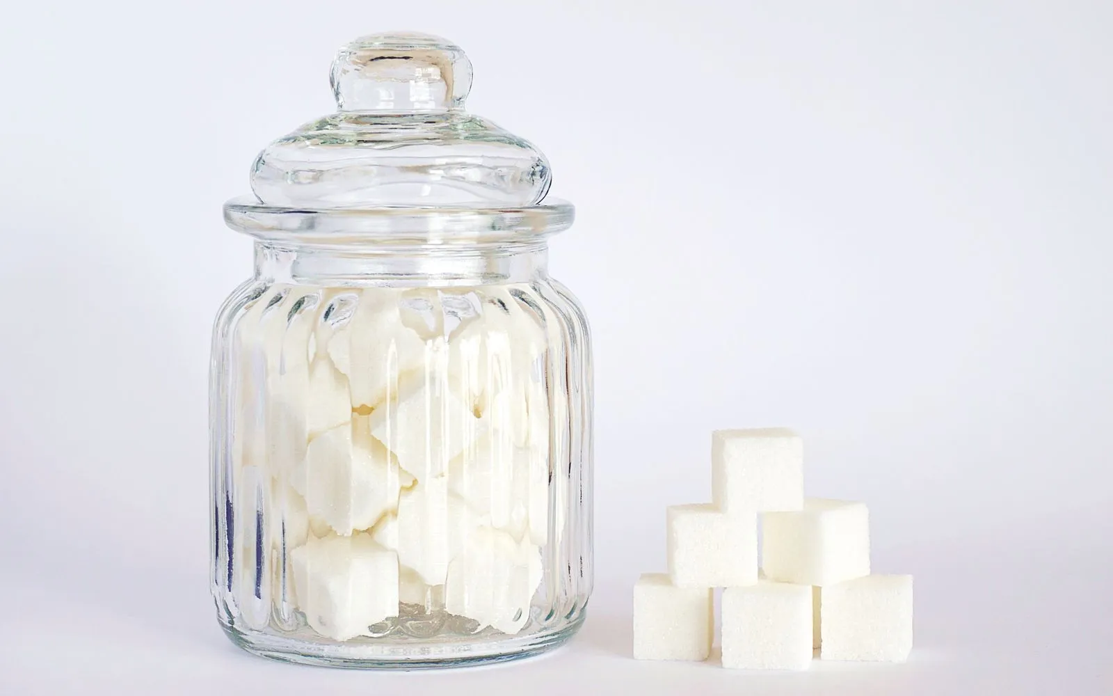 A glass jar of white sugar cubes beside a small stack of cubes on a pale background
