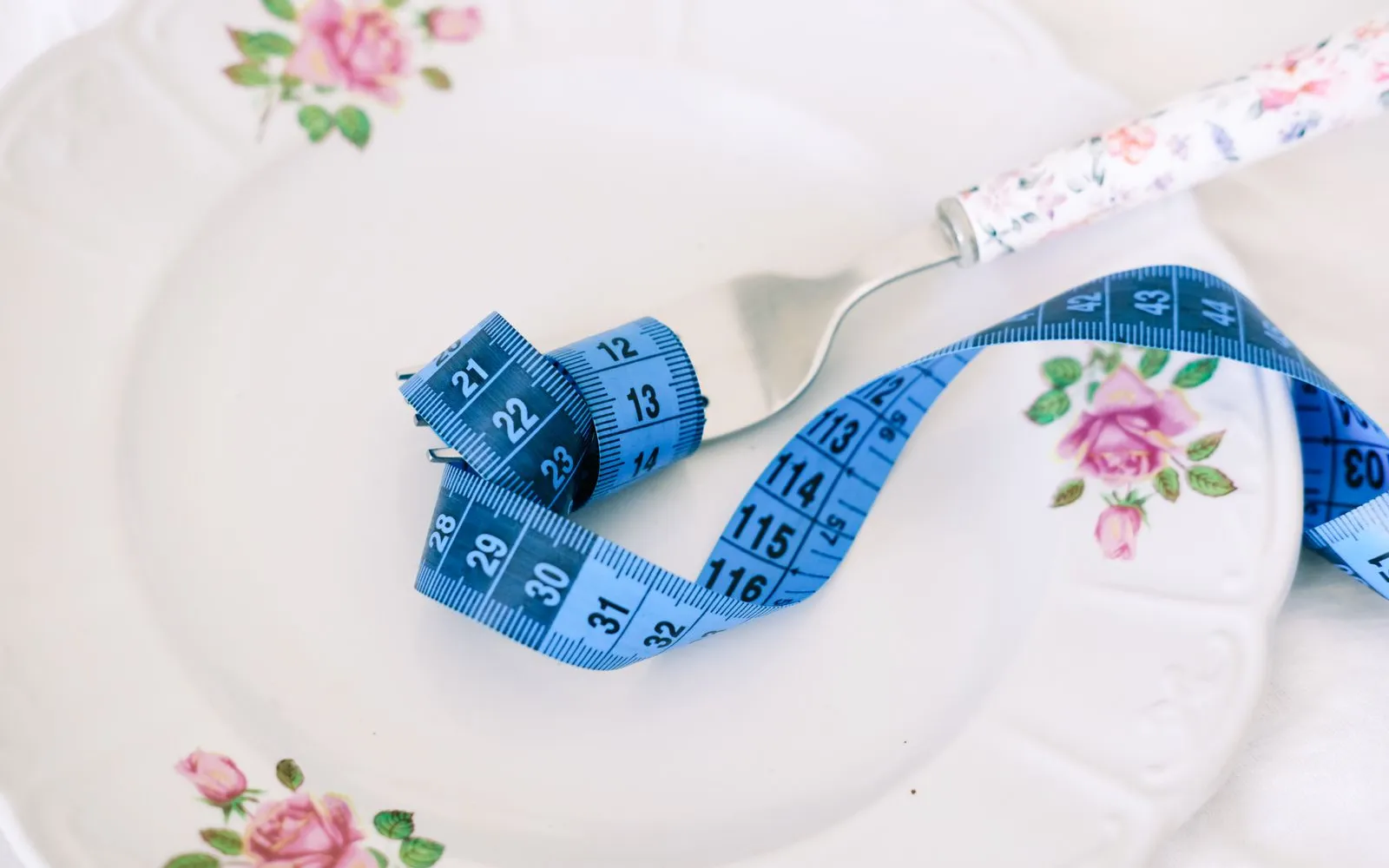 A blue tape measure wrapped around a fork on a white plate with a pink rose pattern.