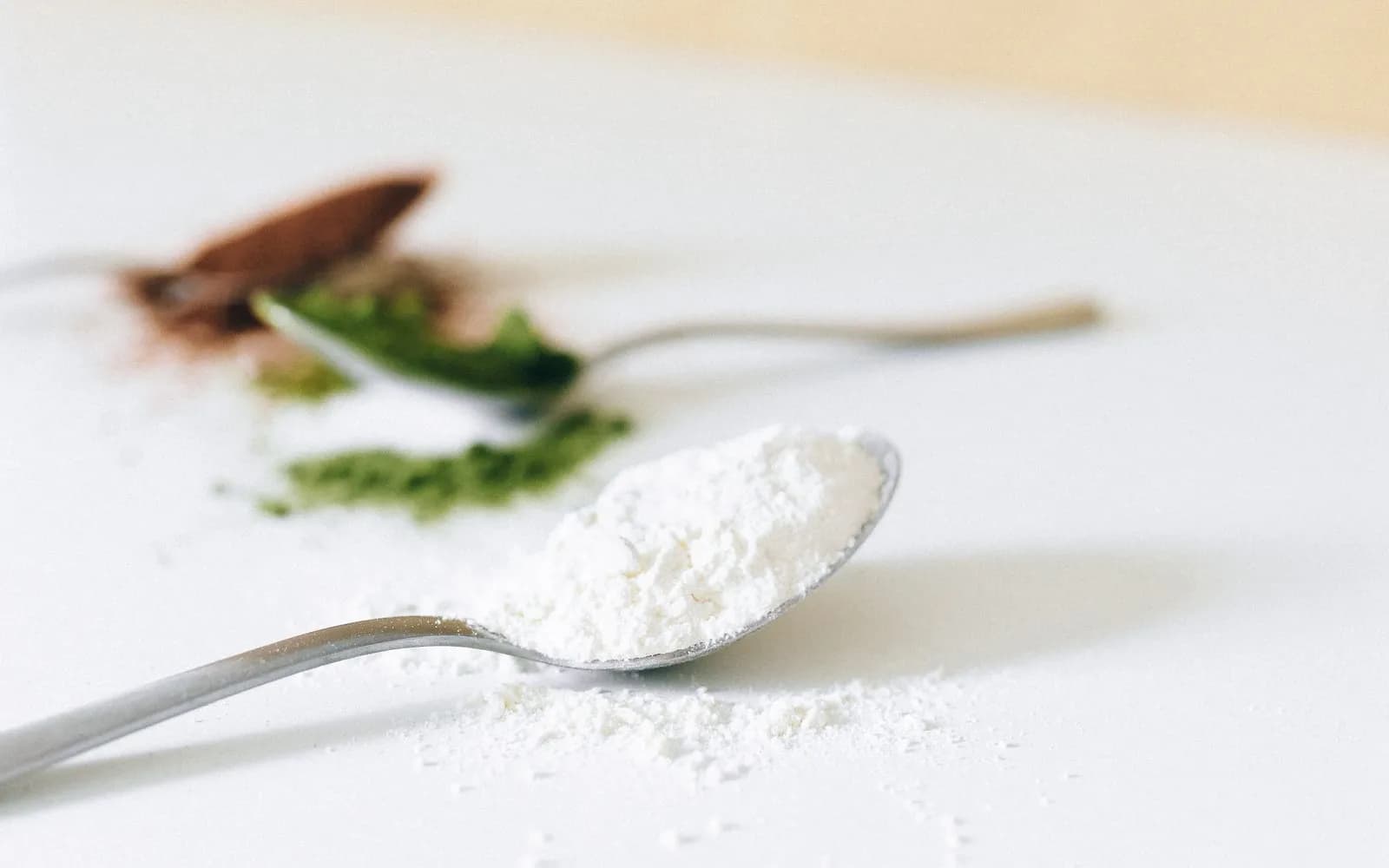 A teaspoon heaped with fine white powder on a white table, with blurred spoons of green and brown powder behind