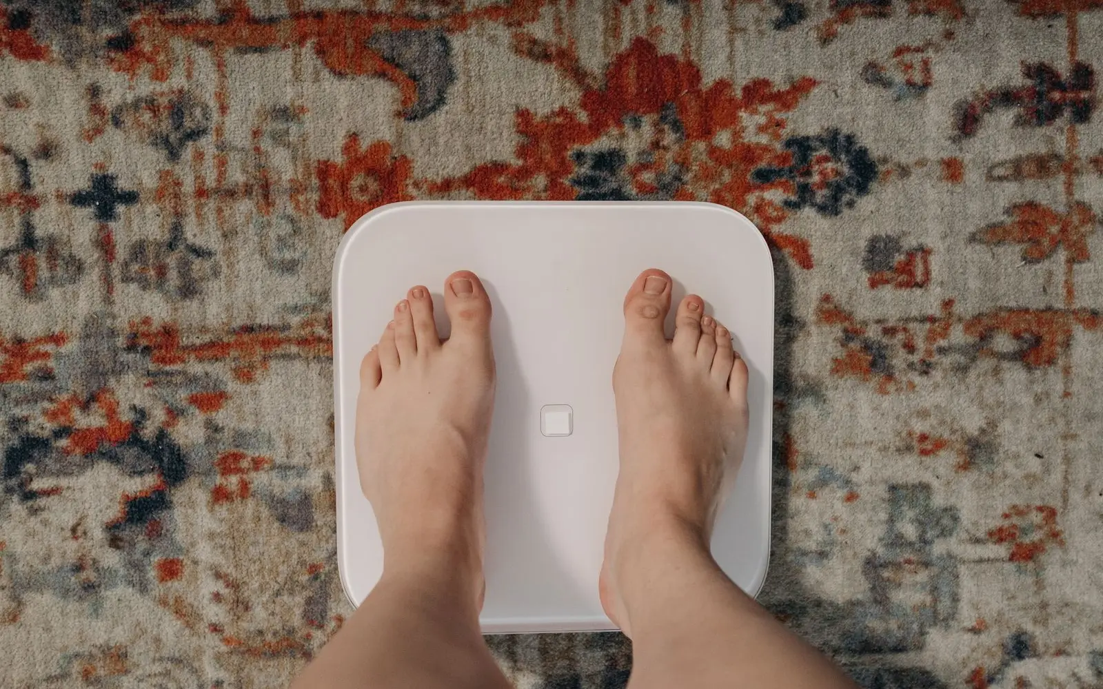 Bare feet standing on a plain white bathroom scale on a patterned rug, seen from above.
