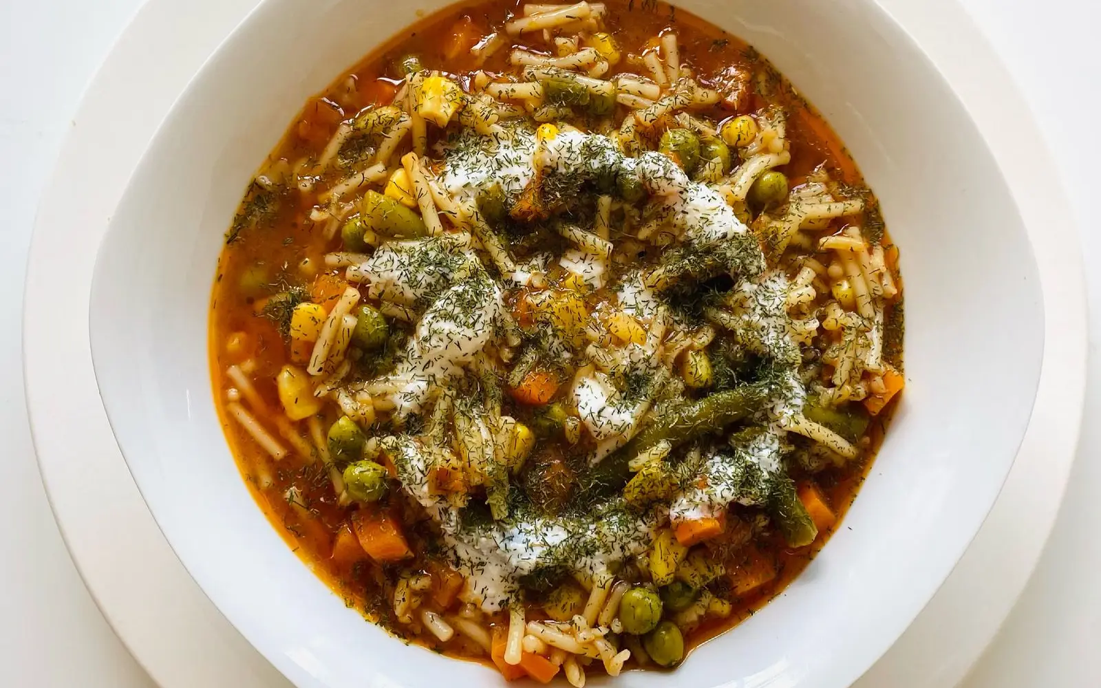 A white bowl of vegetable soup with peas, carrots, green beans, sweetcorn, noodles, dill and a swirl of yoghurt, seen from above.