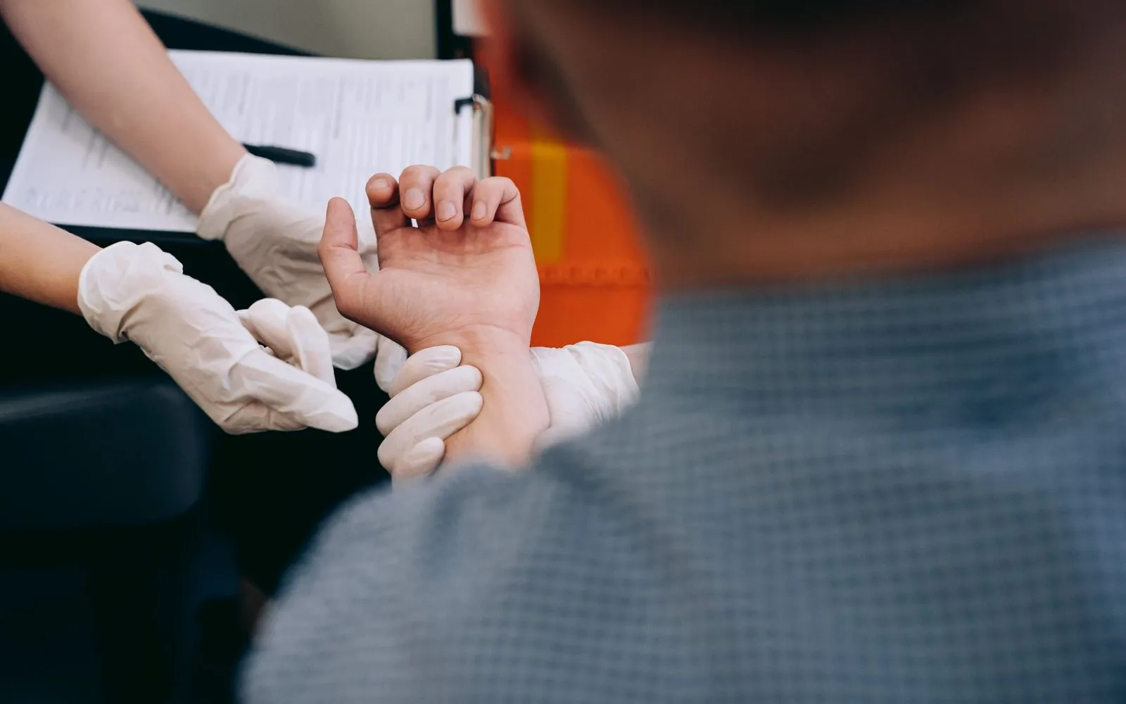 A gloved hand checks a man's pulse at his wrist, with a clipboard in the background