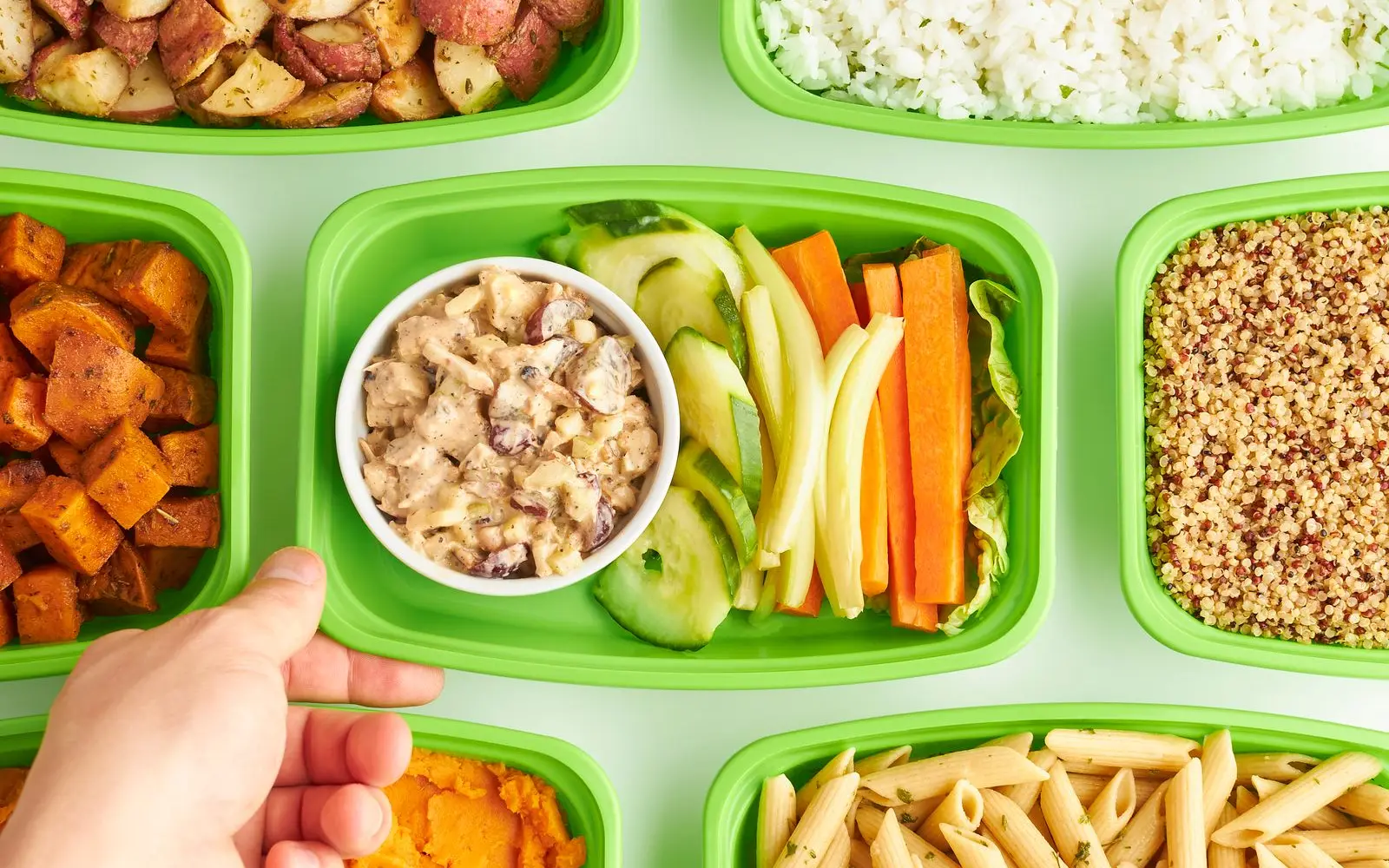 A hand reaching for reusable green meal prep boxes filled with roast potatoes, rice, quinoa, pasta, sweet potato and raw veg.