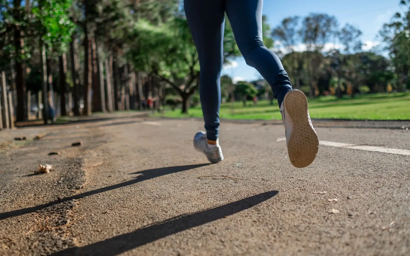 Lower legs of a runner in dark leggings and plain grey trainers, jogging on a park path in the sun.